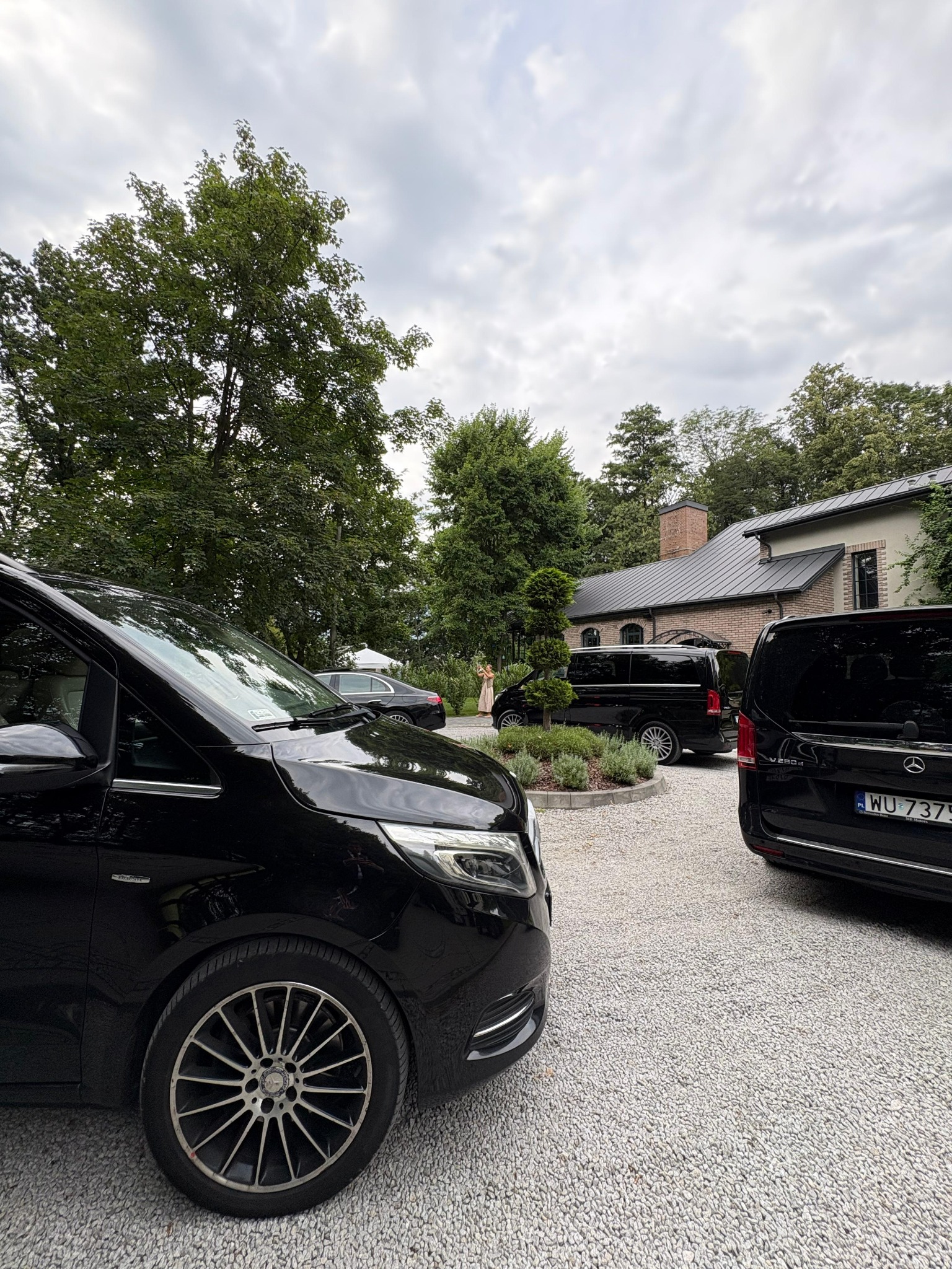 Flota czarnych vanów Mercedes V-klasy na żwirowym podjeździe przed budynkiem. Jeden z vanów z widoczną tablicą rejestracyjną. W tle zieleń drzew i krzewów.