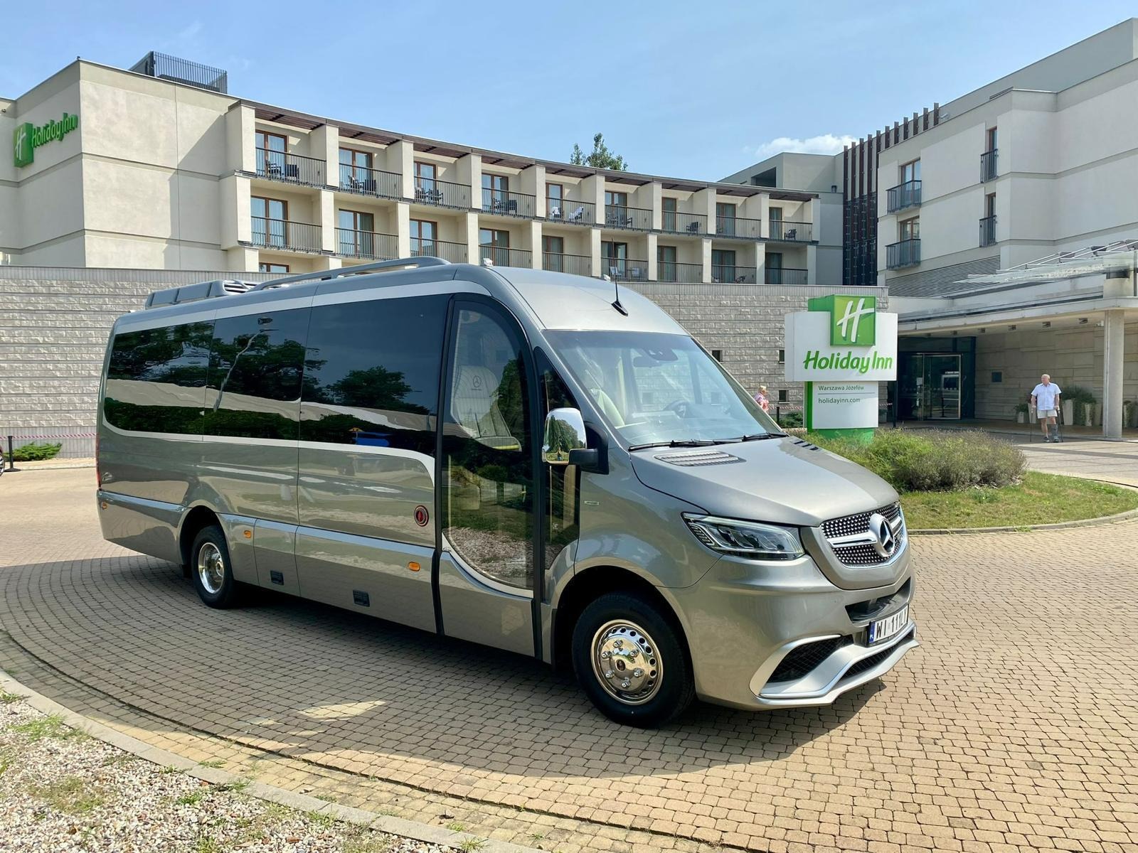 Elegancki, srebrny bus Mercedes-Benz Sprinter z przyciemnianymi szybami, zaparkowany przed hotelem Holiday Inn w słoneczny dzień. Widoczny fragment budynku i logo hotelu.