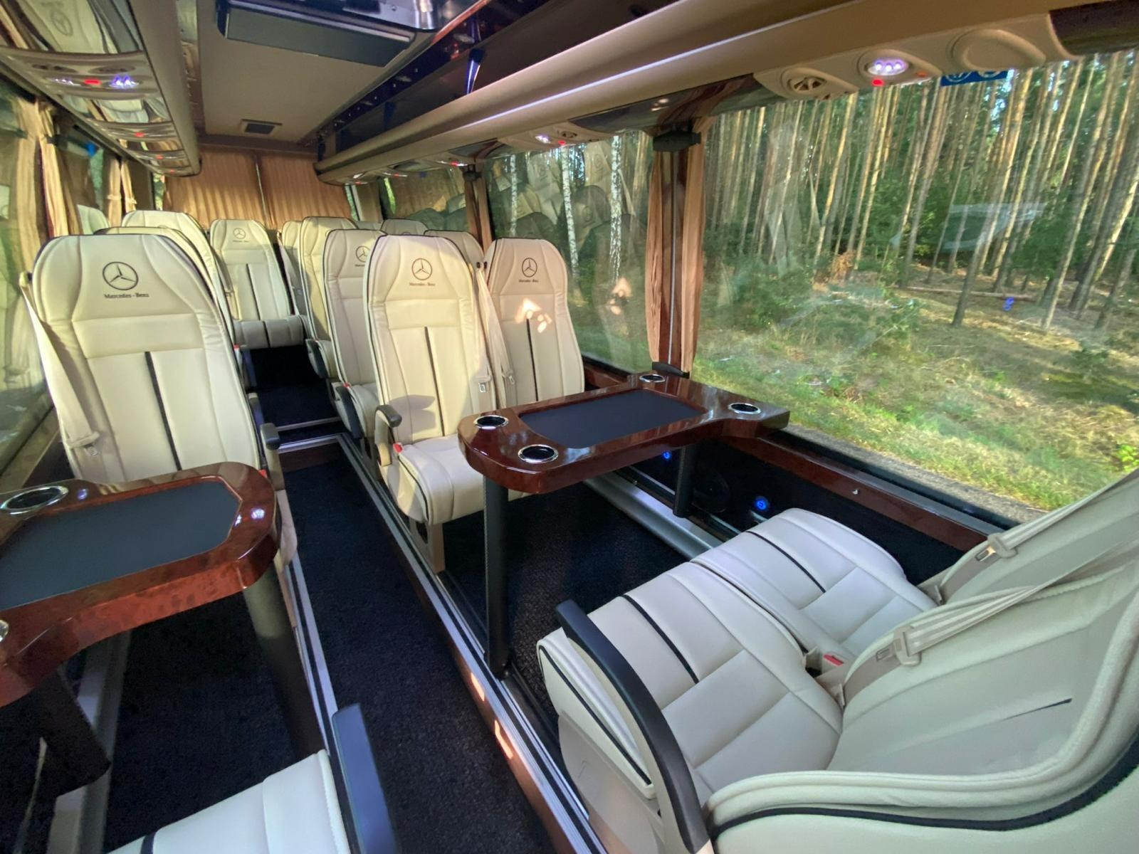 Luksusowe wnętrze busa Mercedes-Benz z beżowymi skórzanymi fotelami, drewnianymi stolikami i widokiem na las za oknem. Komfortowe podróżowanie.