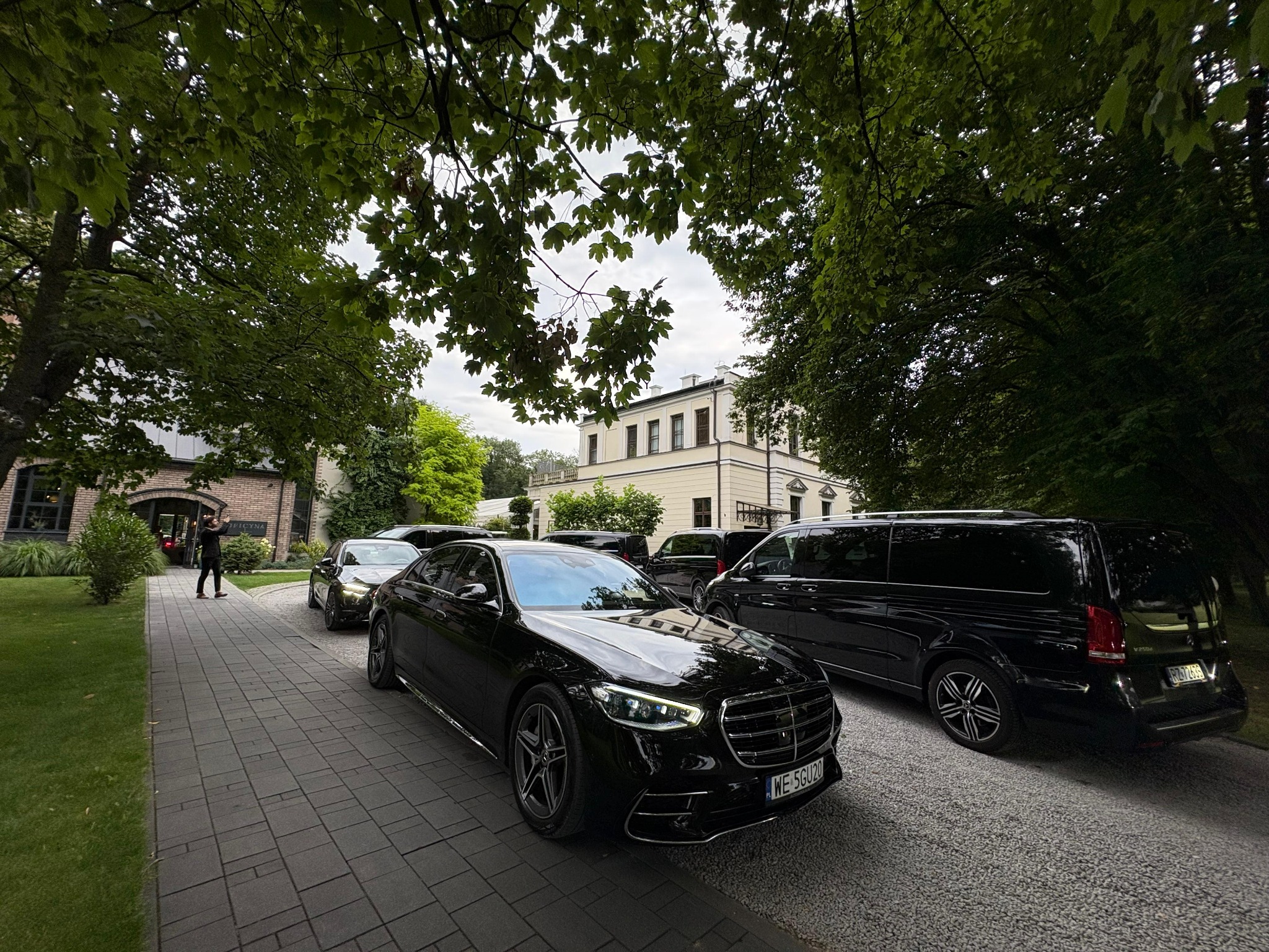 Flota luksusowych, czarnych samochodów (Mercedes S-klasa i V-klasa) zaparkowana przed budynkiem, otoczona zielenią. Osoba w tle. Elegancki transport.