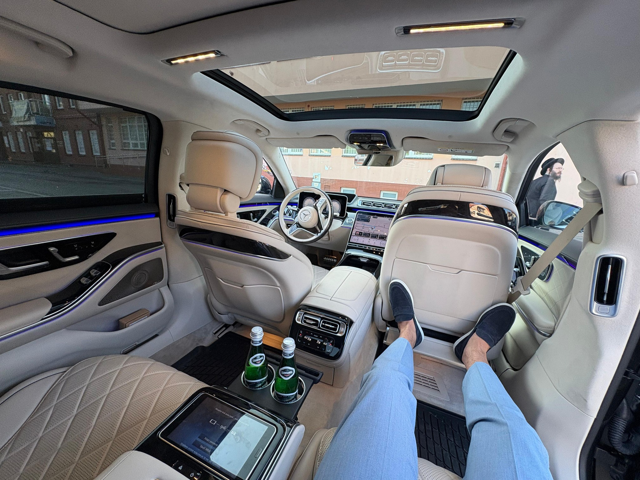 Luksusowe wnętrze Mercedesa klasy S z jasną skórzaną tapicerką, panoramicznym dachem i widocznymi nogami pasażera z tyłu. Dwie butelki wody mineralnej w uchwycie.