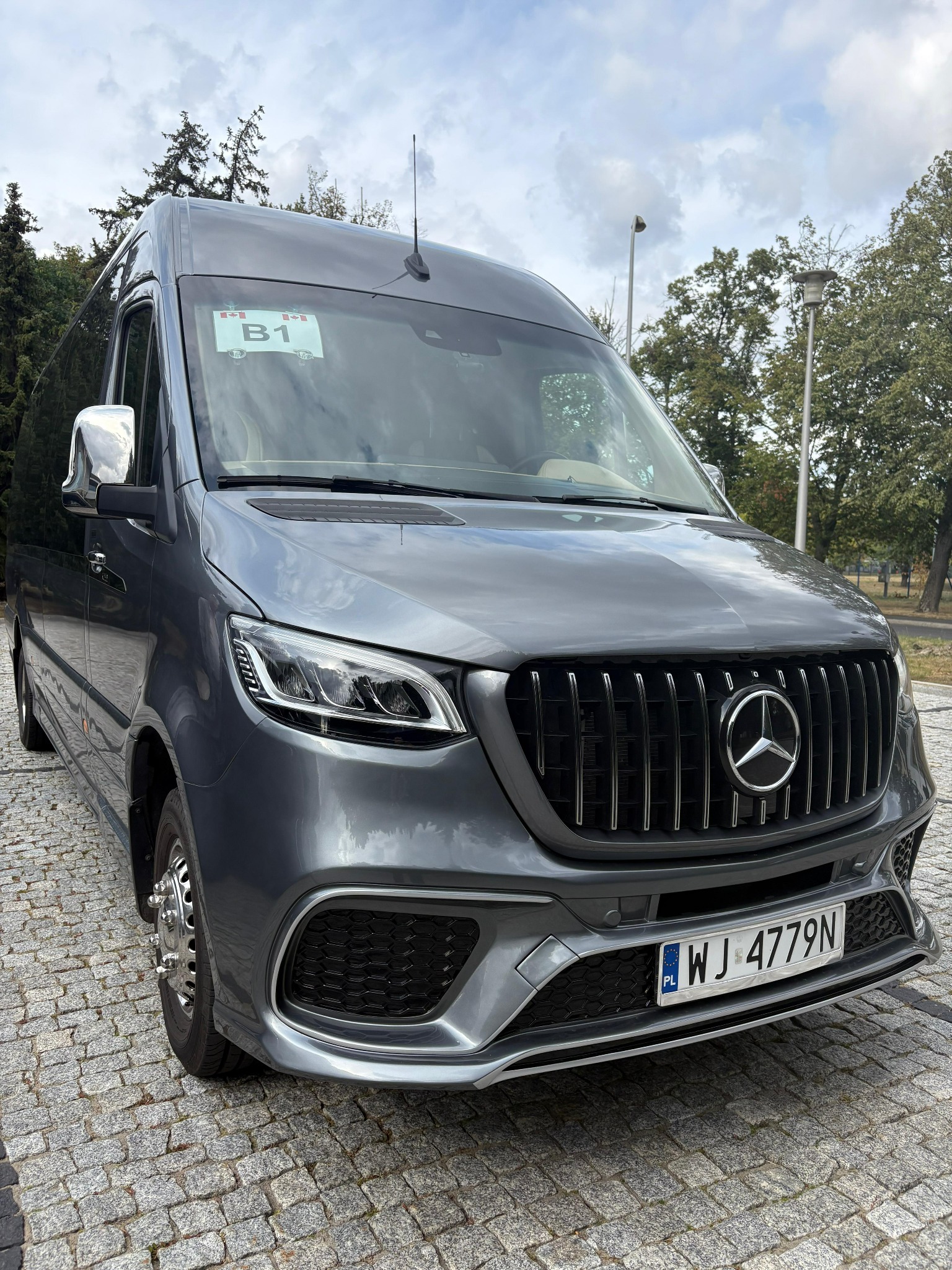 Szary, luksusowy bus Mercedes-Benz Sprinter z chromowanymi lusterkami i charakterystycznym grillem AMG, zaparkowany na brukowanej nawierzchni w słoneczny dzień.