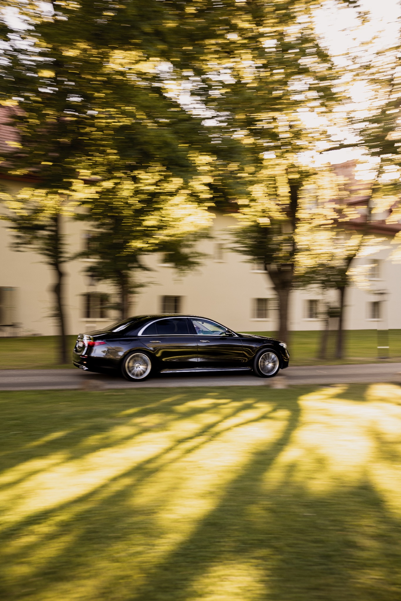 Czarna limuzyna w ruchu, rozmyte tło drzew i budynków, dynamiczne ujęcie z boku, promienie słoneczne przebijające przez korony drzew.