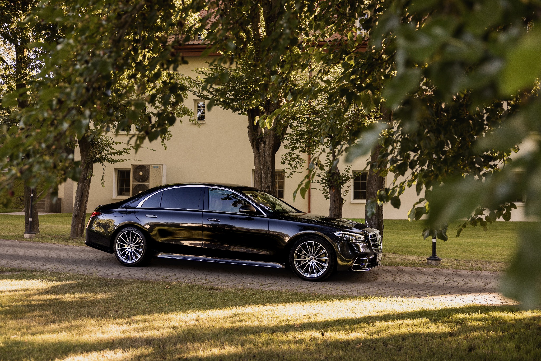 Elegancka, czarna limuzyna Mercedes-Benz S-Class, zaparkowana na brukowanej drodze w otoczeniu zieleni i drzew, z budynkiem w tle. Luksusowy transport.