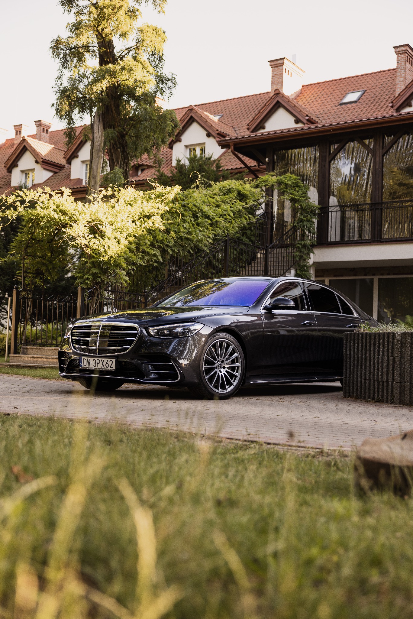 Elegancki, czarny Mercedes klasy S, zaparkowany na podjeździe luksusowej rezydencji z czerwonym dachem i bujną zielenią. Widoczne detale: chromowana atrapa chłodnicy.