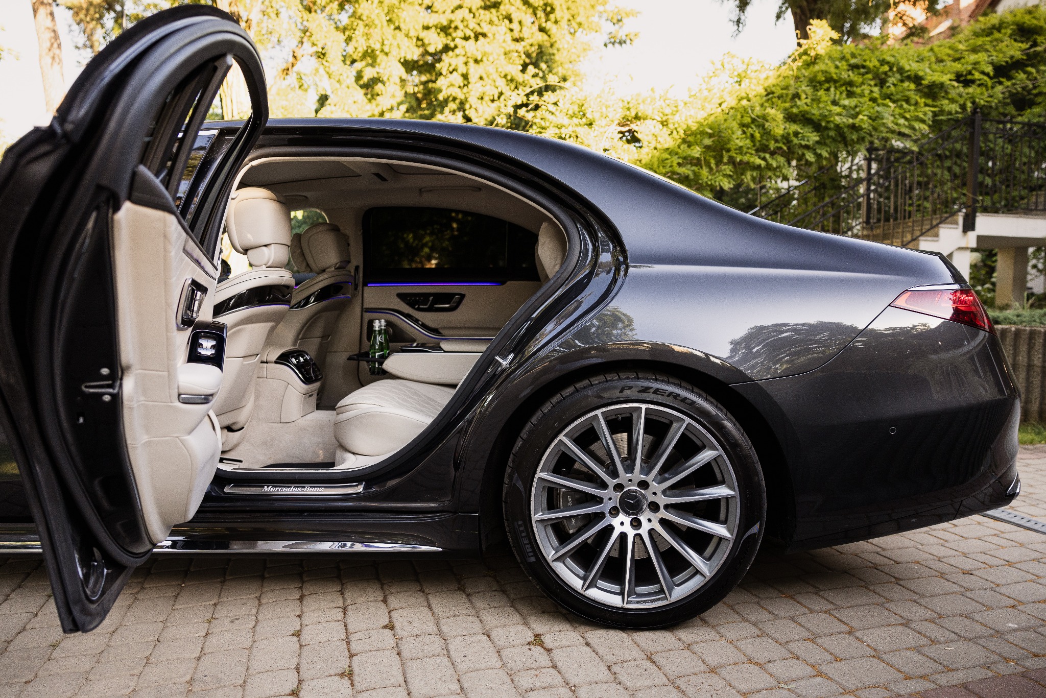 Luksusowy, grafitowy Mercedes-Benz S-klasa z otwartymi drzwiami, ukazujący kremowe skórzane wnętrze z subtelnym oświetleniem ambientowym i butelką wody na tylnym siedzeniu.