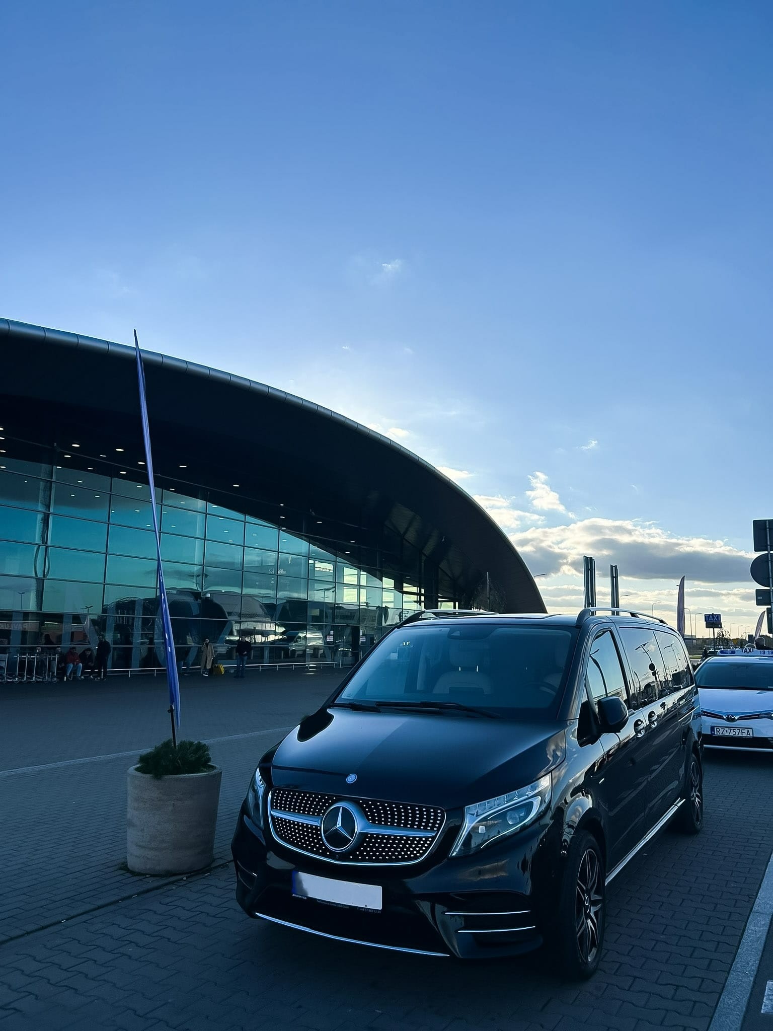 Czarny Mercedes V-klasa na tle nowoczesnego budynku lotniska w słoneczny dzień. Widoczny fragment chodnika z kostki brukowej i inne pojazdy.