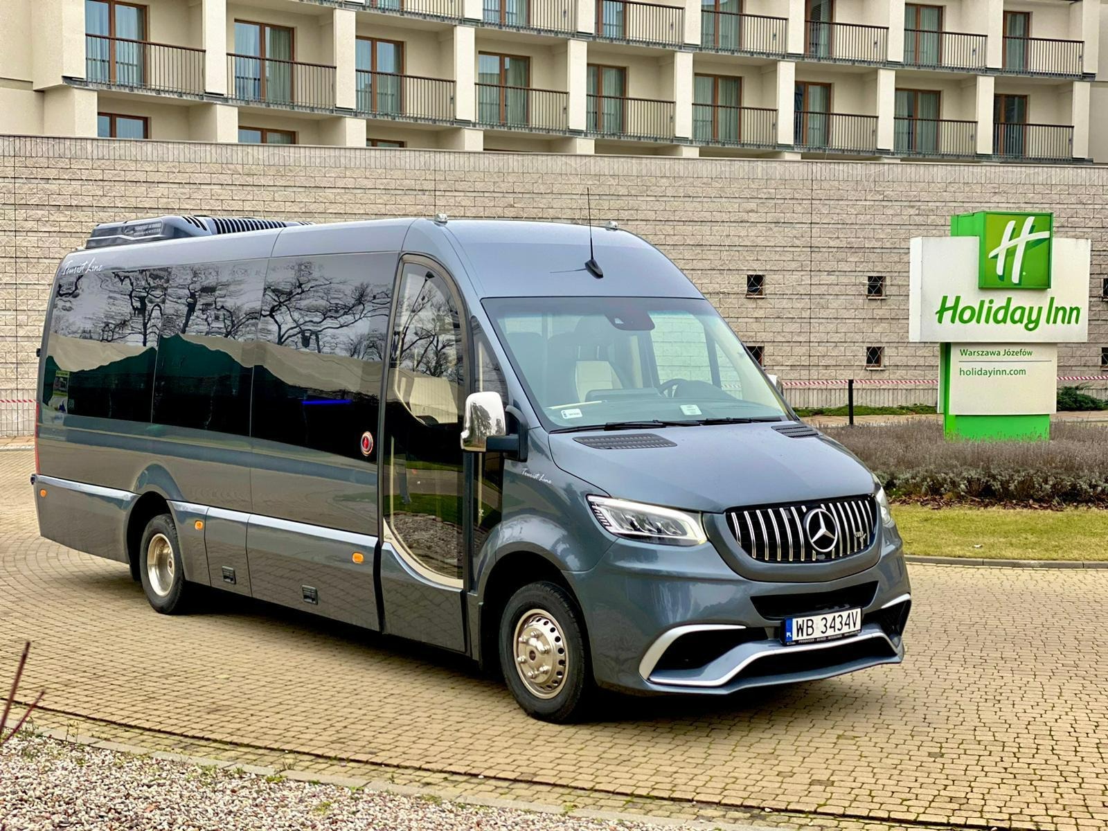 Szary, luksusowy bus Mercedes-Benz z otwartymi drzwiami na tle hotelu Holiday Inn. Widoczne odbicie drzew w szybach. Tablica rejestracyjna WB 3434V.