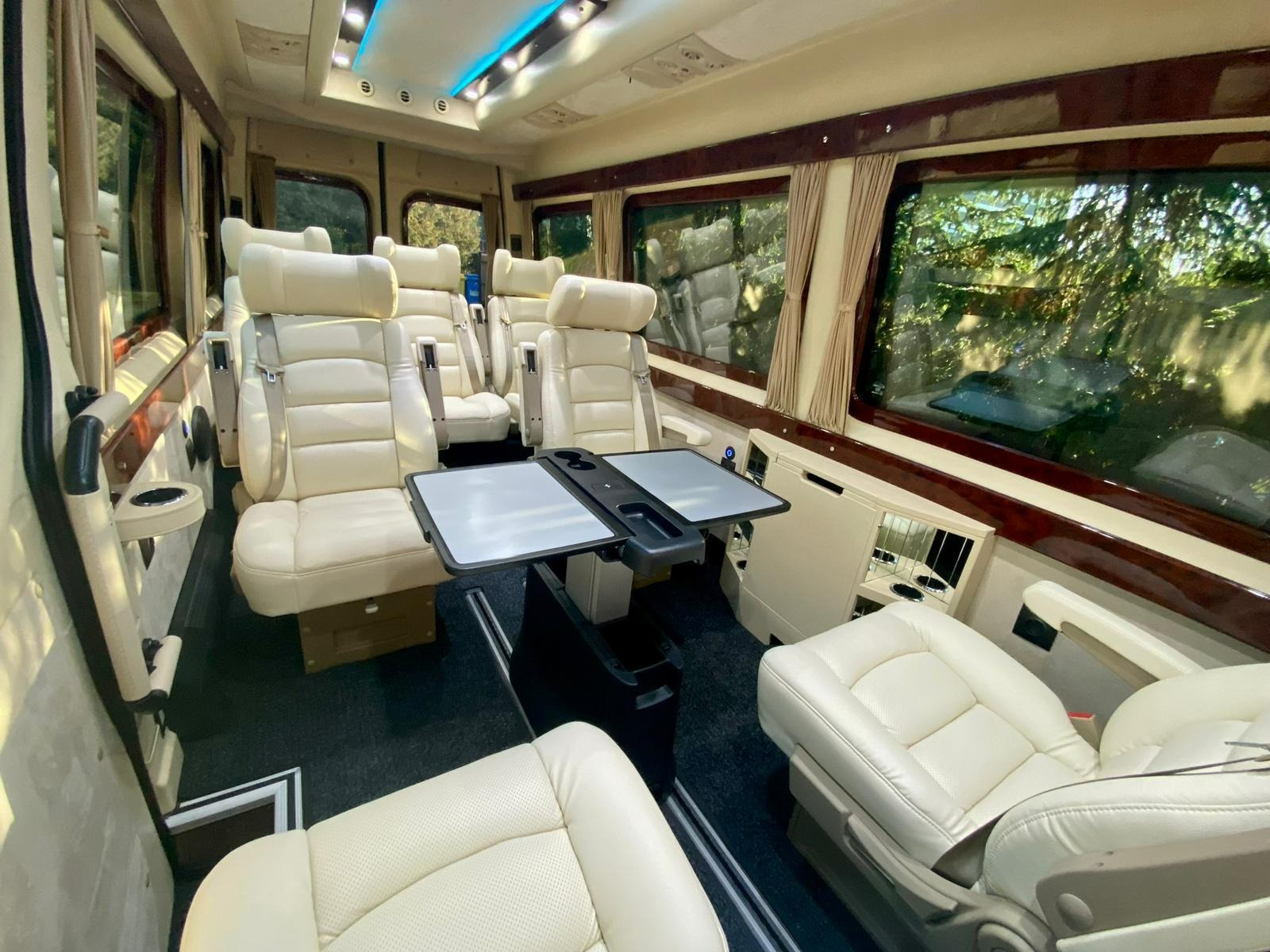 Luksusowe wnętrze busa z kremowymi skórzanymi fotelami, stolikami i oknami z zasłonami. Komfortowy transport VIP z oświetleniem LED.