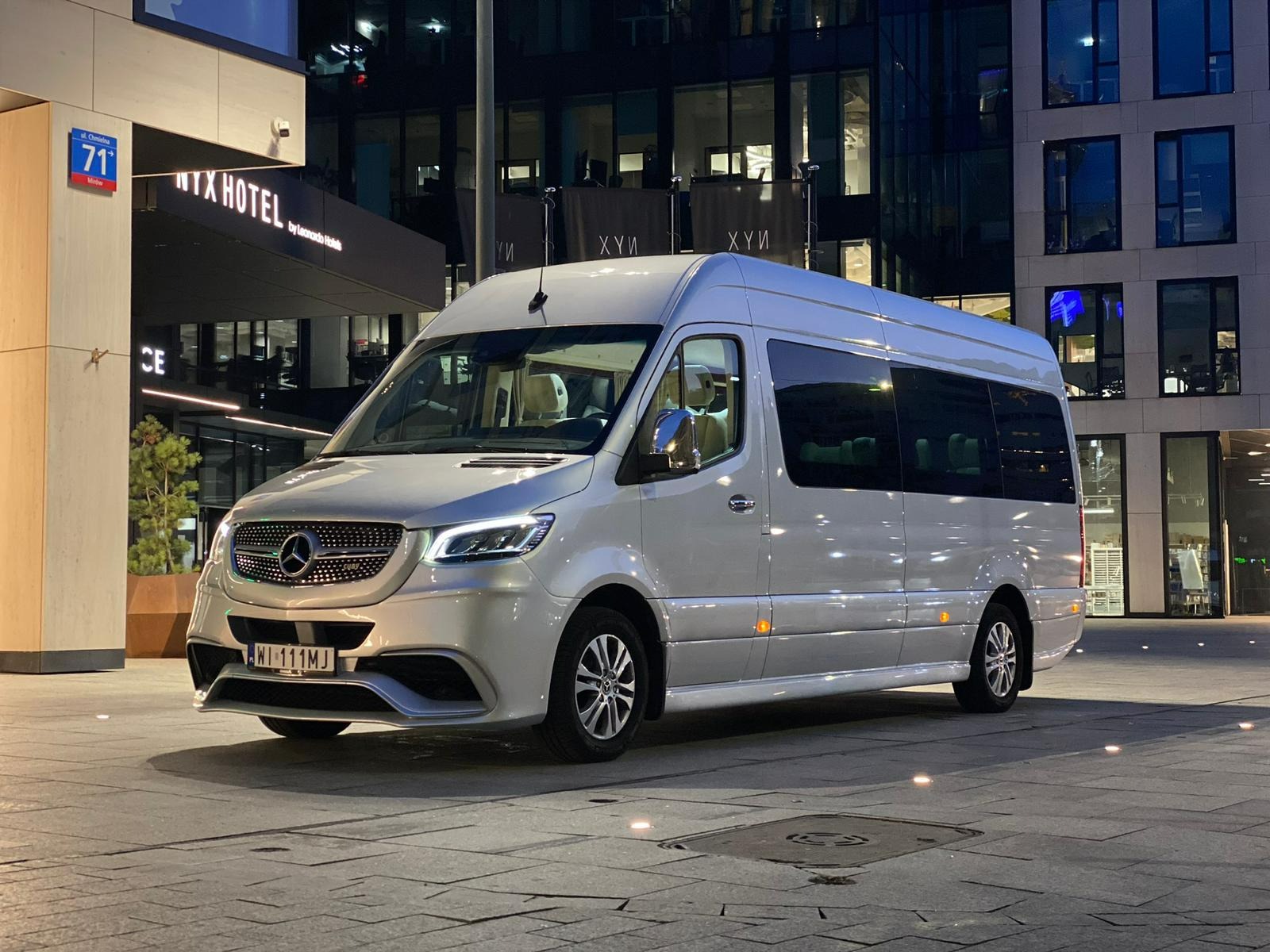 Srebrny Mercedes Sprinter z przyciemnianymi szybami, zaparkowany nocą przed nowoczesnym budynkiem z napisem 'NYX HOTEL'. Widoczny znak uliczny 'ul. Chmielna 71'.