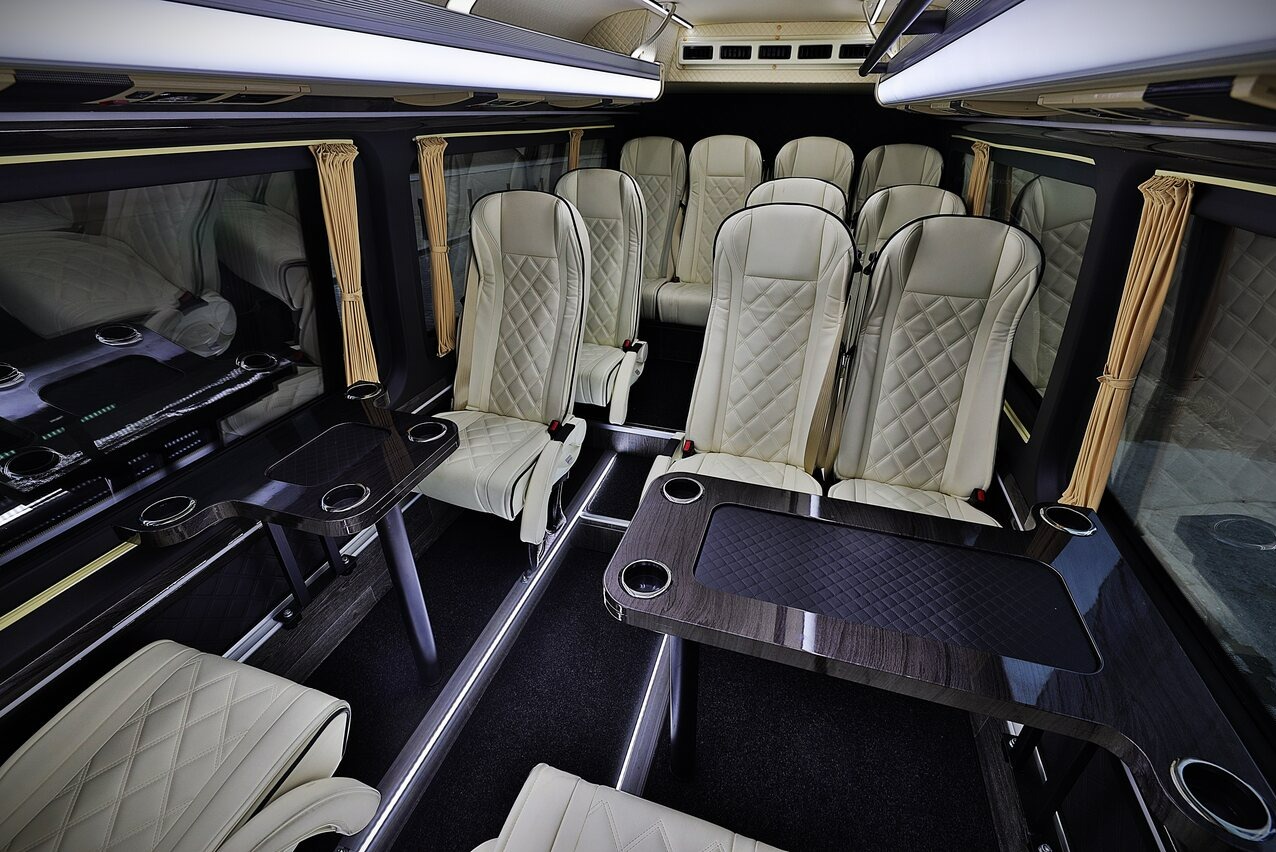 Luksusowe wnętrze busa z kremowymi, pikowanymi fotelami, drewnianymi stolikami z uchwytami na napoje i oświetleniem LED w podłodze. Eleganckie zasłony w oknach.
