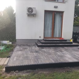 LevelStone Mykhailo Habrus - Nowoczesny taras z kostki brukowej w odcieniach szarości, z niskimi schodami prowadzącymi do domu. Widoczny fragment trawnika i roślinność w tle.