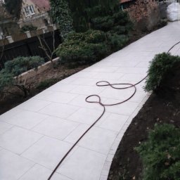 LevelStone Mykhailo Habrus - Jasna, szeroka ścieżka z płyt brukowych w ogrodzie, wijąca się między zielenią. W tle widoczny dom z czerwonym dachem i fragment czarnego ogrodzenia. Wąż ogrodowy leży na ścieżce.