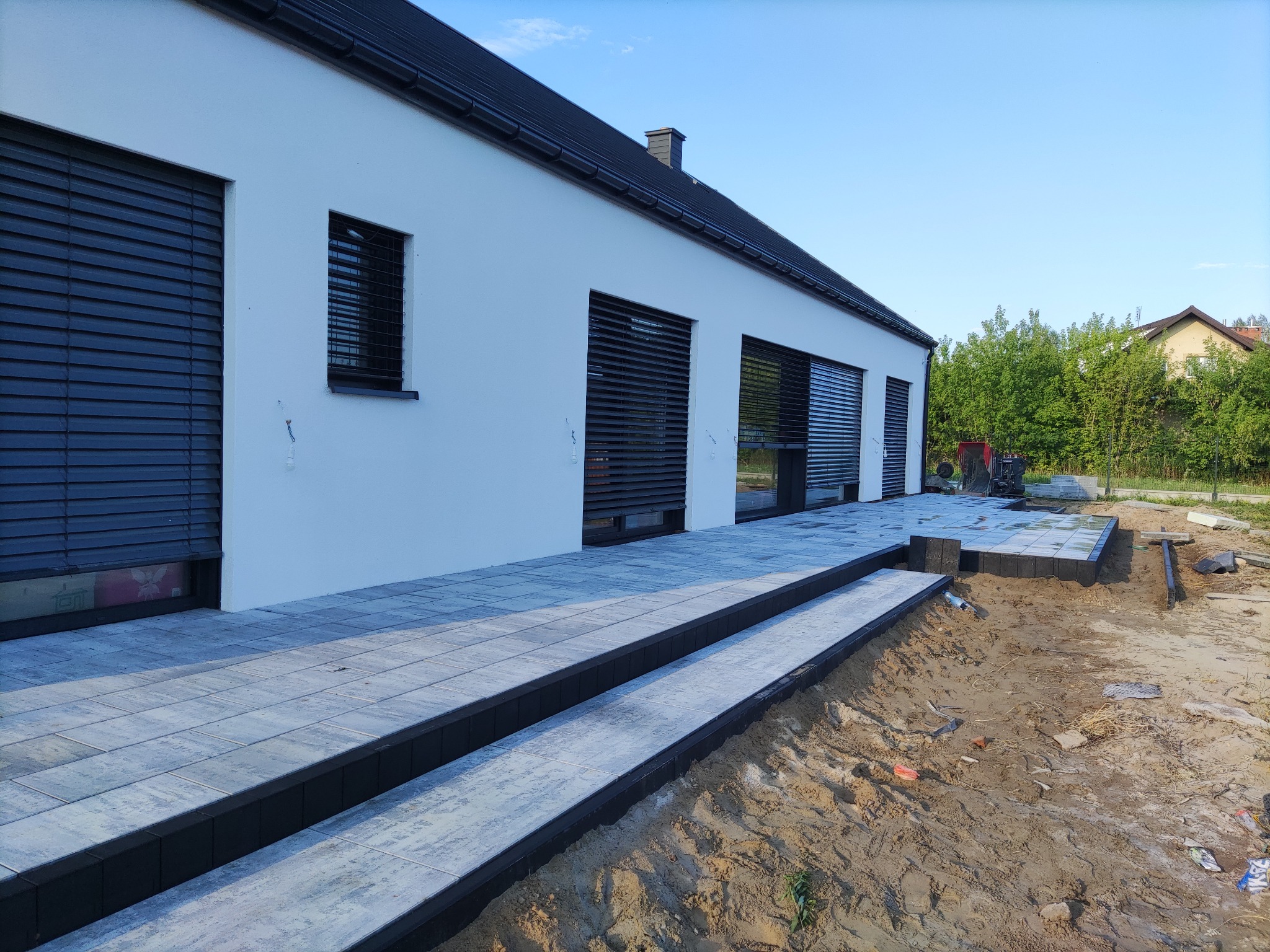 Nowoczesny dom z szarą kostką brukową na tarasie i schodach. W tle widoczny teren przygotowany pod dalsze prace brukarskie i maszyna budowlana.