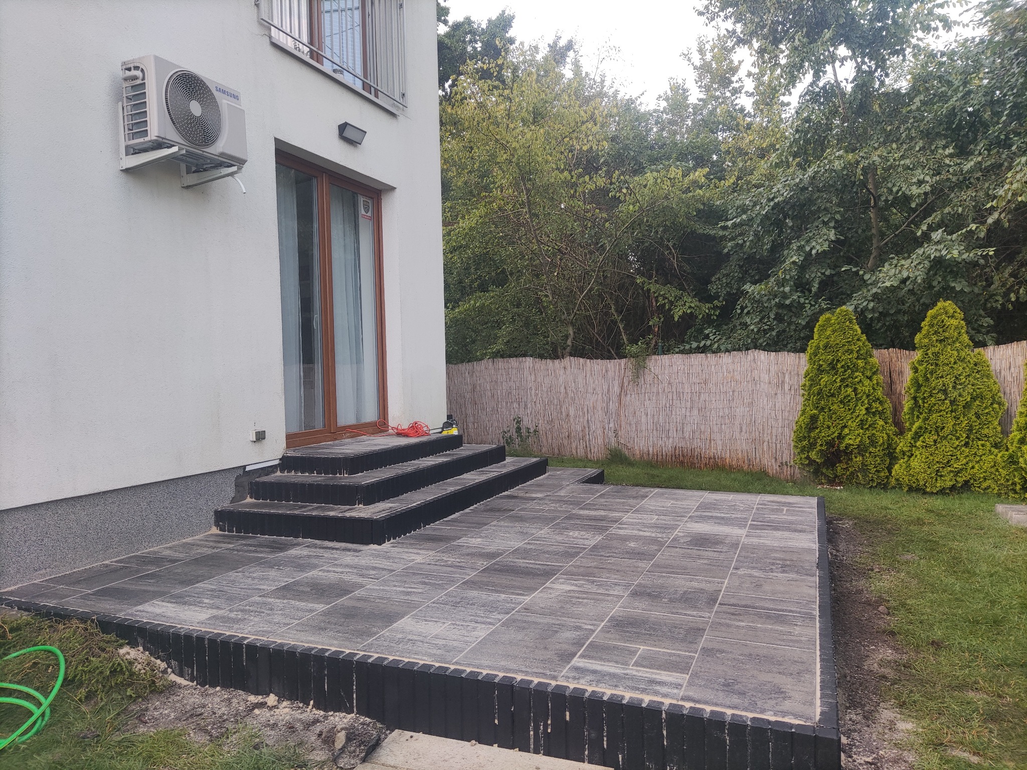 Ułożona kostka brukowa w odcieniach szarości tworząca taras z schodami przy domu. Obramowanie z ciemnych, pionowo ułożonych elementów. Widok na ogród i roślinność.