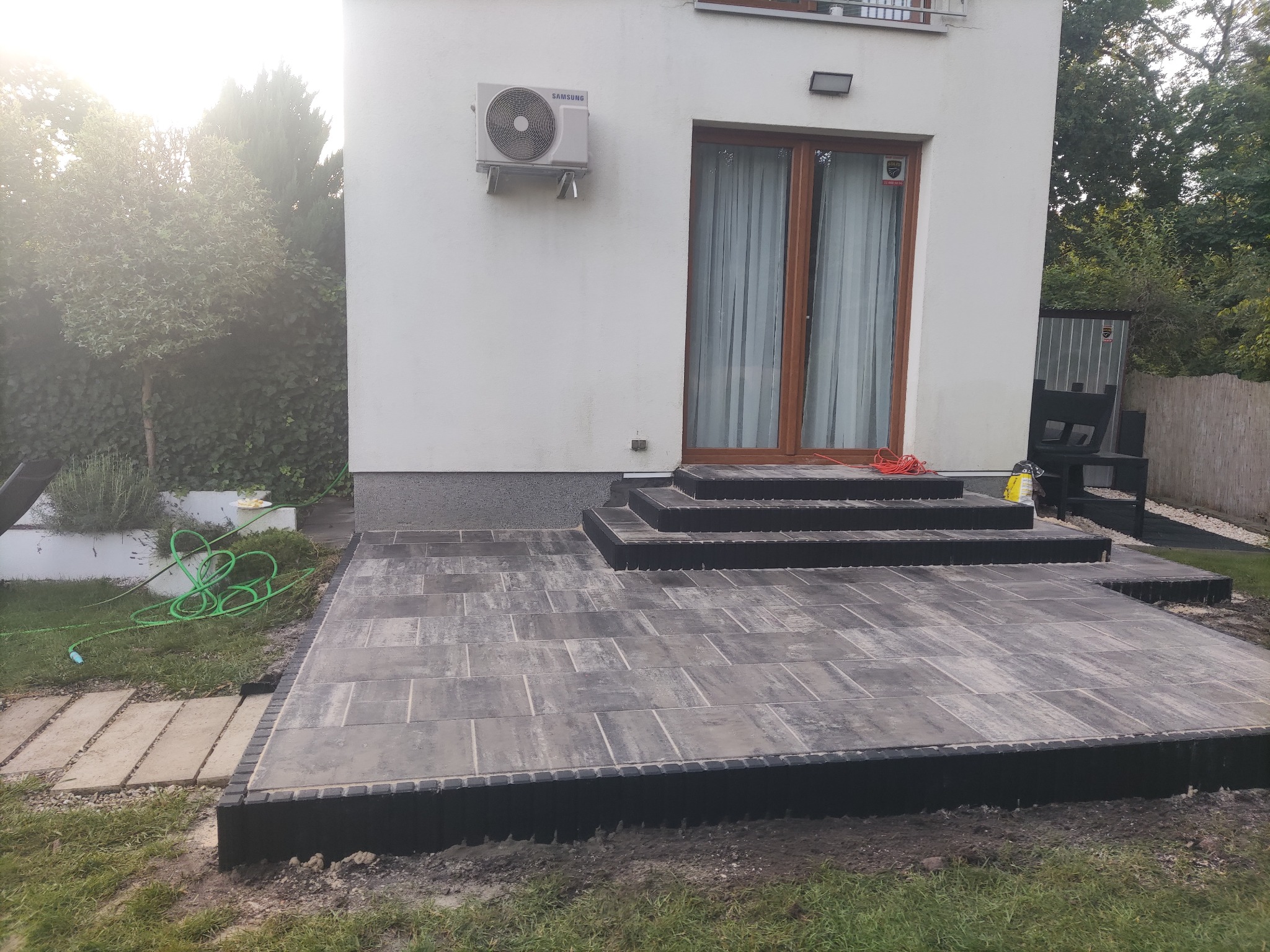 Nowoczesny taras z kostki brukowej w odcieniach szarości, z niskimi schodami prowadzącymi do domu. Widoczny fragment trawnika i roślinność w tle.
