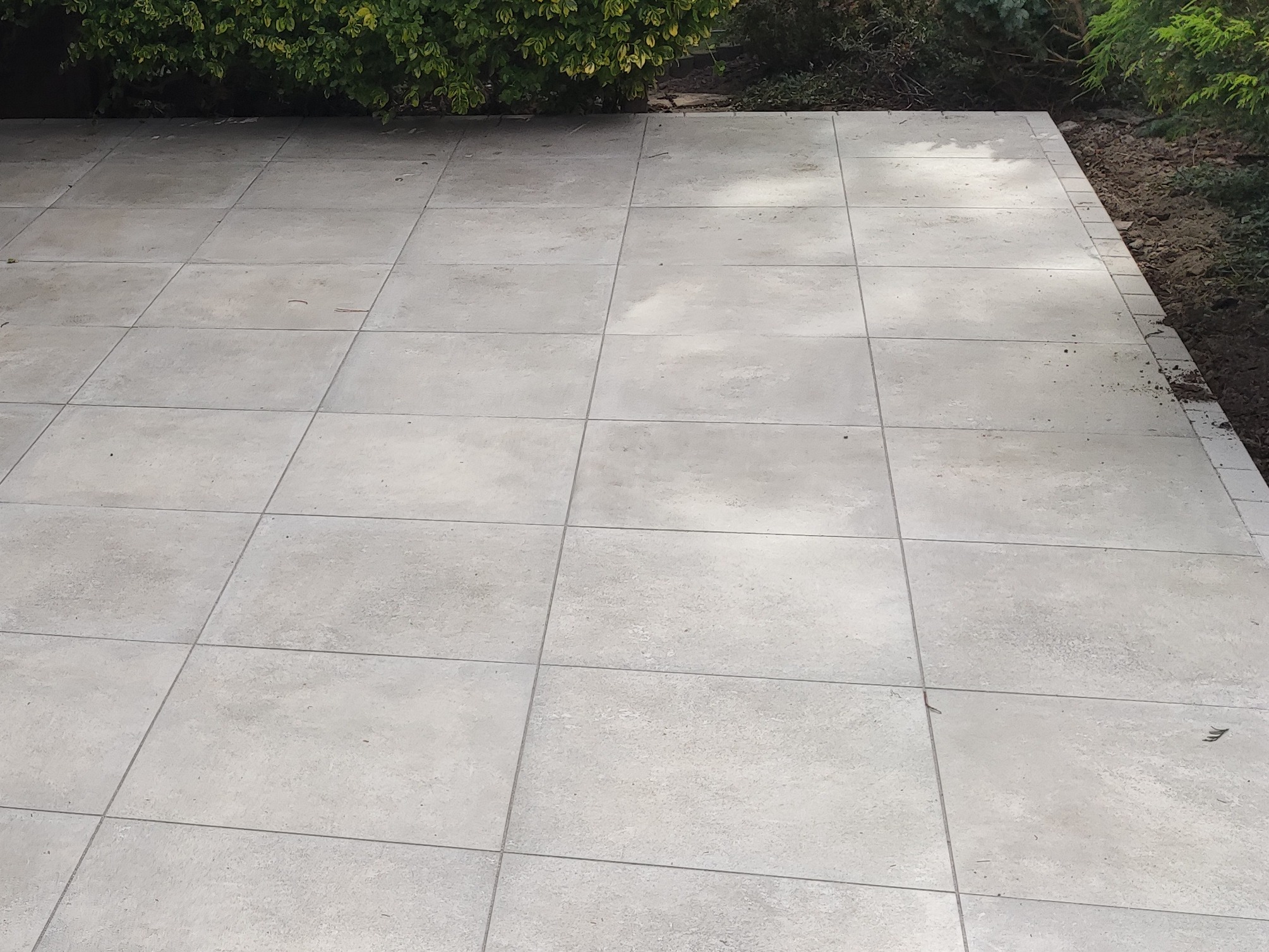 Jasnoszara, regularna kostka brukowa ułożona na tarasie, z widoczną roślinnością w tle. Minimalistyczny design i precyzyjne wykonanie.