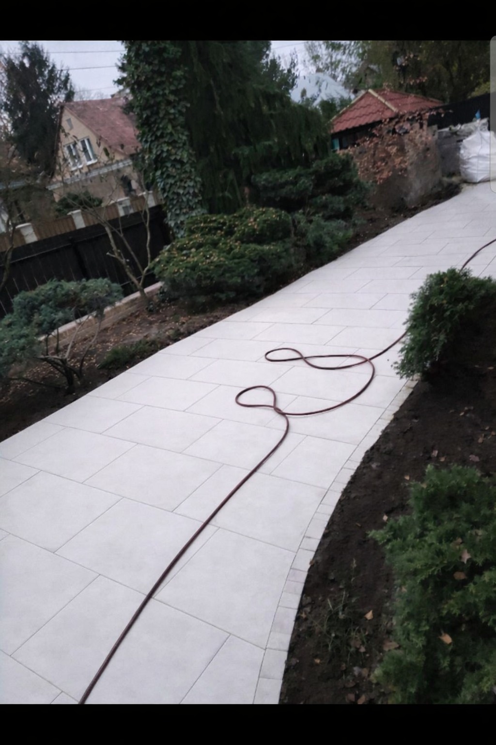 Jasna, szeroka ścieżka z płyt brukowych w ogrodzie, wijąca się między zielenią. W tle widoczny dom z czerwonym dachem i fragment czarnego ogrodzenia. Wąż ogrodowy leży na ścieżce.