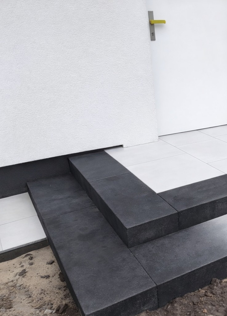 Nowoczesne schody wejściowe z ciemnej kostki brukowej, kontrastujące z białą elewacją i płytkami. Minimalistyczny design, precyzyjne wykonanie.