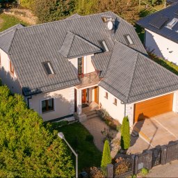 Wrona nieruchomości - Widok z góry na dom jednorodzinny z szarą dachówką, brązową bramą garażową i zadbanym ogrodem. Na dachu widoczne okna połaciowe i antena satelitarna.