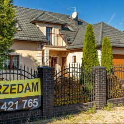 Wrona nieruchomości - Dom jednorodzinny z ogłoszeniem 'Sprzedam' na ogrodzeniu z czarnej cegły. Widoczne tuje i brama garażowa. Dach pokryty szarą dachówką.
