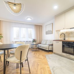 Wrona nieruchomości - Jasne wnętrze mieszkania: stół z dekoracją, krzesła, sofa i zabudowa kuchenna z piekarnikiem. Parkiet w jodełkę, nowoczesna lampa wisząca.
