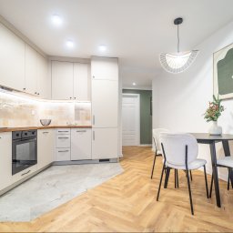 Wrona nieruchomości - Jasna kuchnia z drewnianym blatem i parkietem w jodełkę, okrągły stół z krzesłami i dekoracją świąteczną, nowoczesne oświetlenie i minimalistyczny obraz na ścianie.