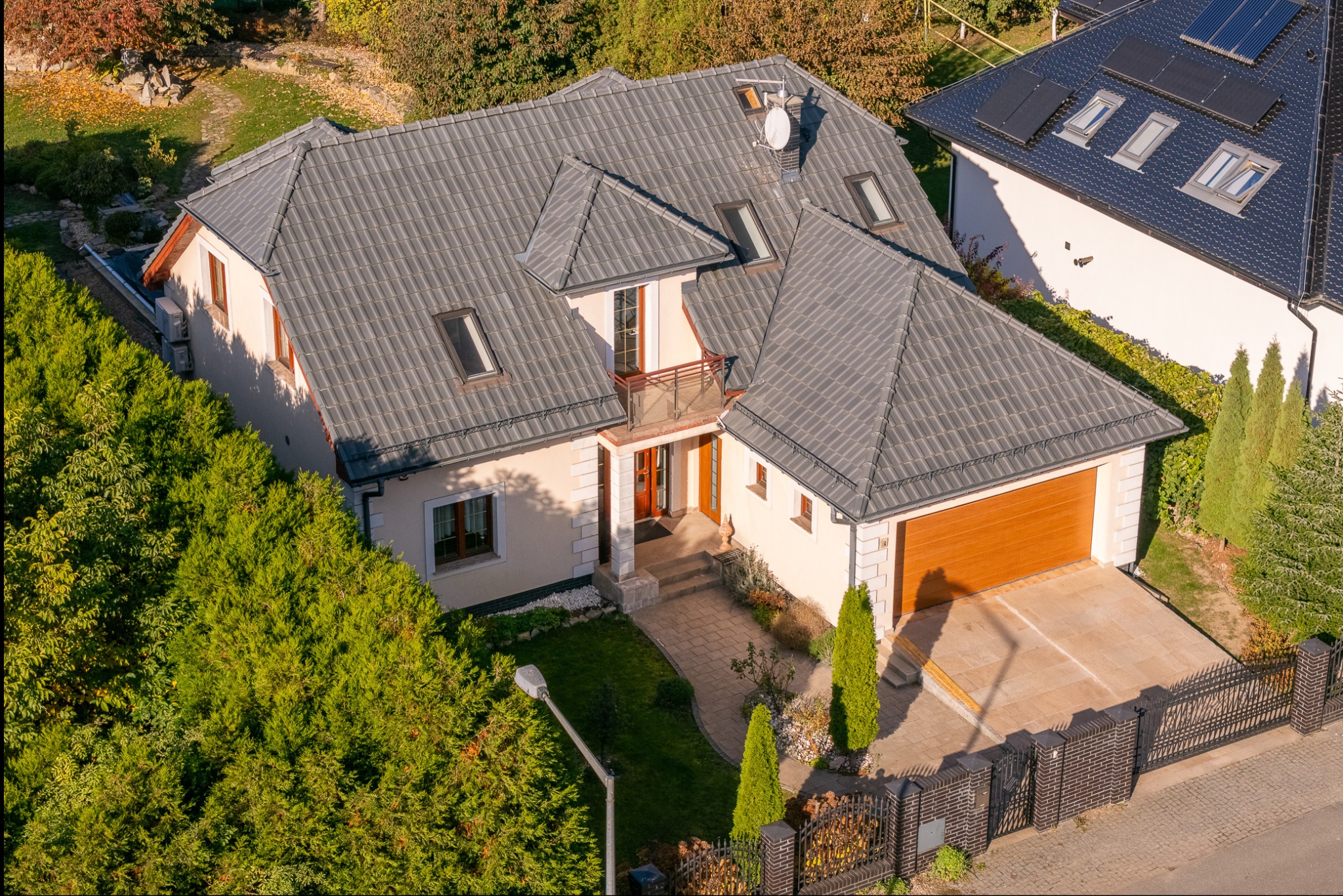 Widok z góry na dom jednorodzinny z szarą dachówką, brązową bramą garażową i zadbanym ogrodem. Na dachu widoczne okna połaciowe i antena satelitarna.