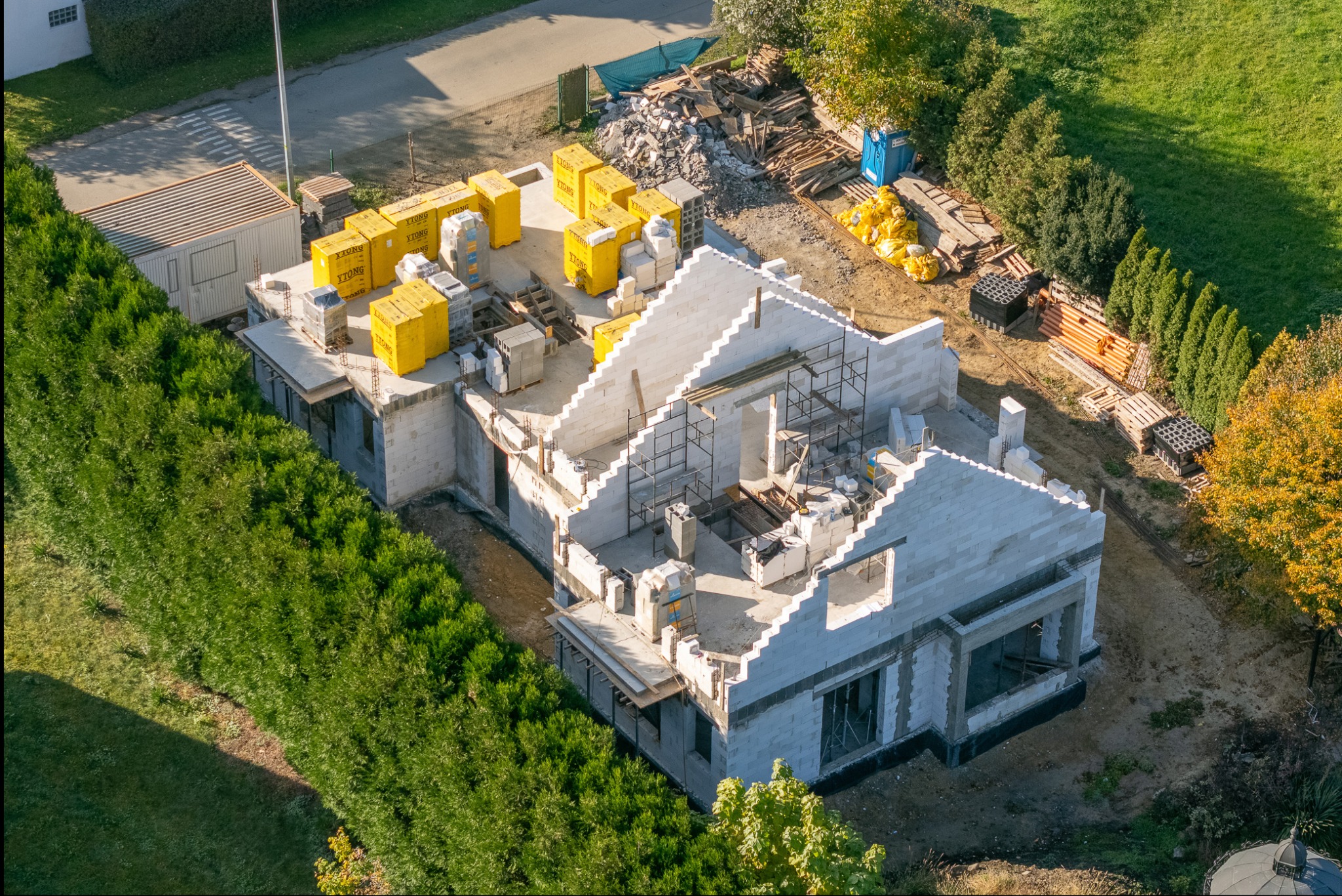 Budowa domu jednorodzinnego z białych bloczków Ytong, widok z góry. Częściowo wzniesione ściany, materiały budowlane na dachu, rusztowania. Otoczenie: zieleń, drzewa.