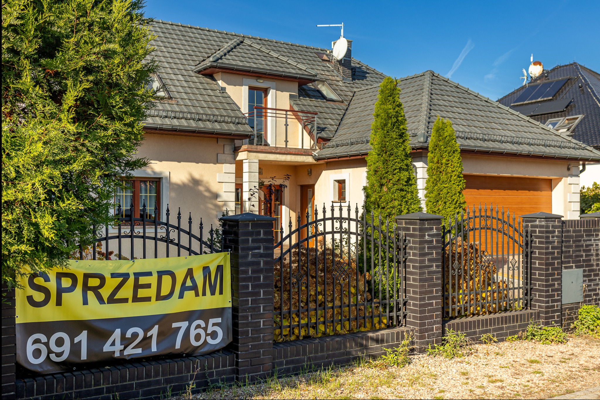 Dom jednorodzinny z ogłoszeniem 'Sprzedam' na ogrodzeniu z czarnej cegły. Widoczne tuje i brama garażowa. Dach pokryty szarą dachówką.