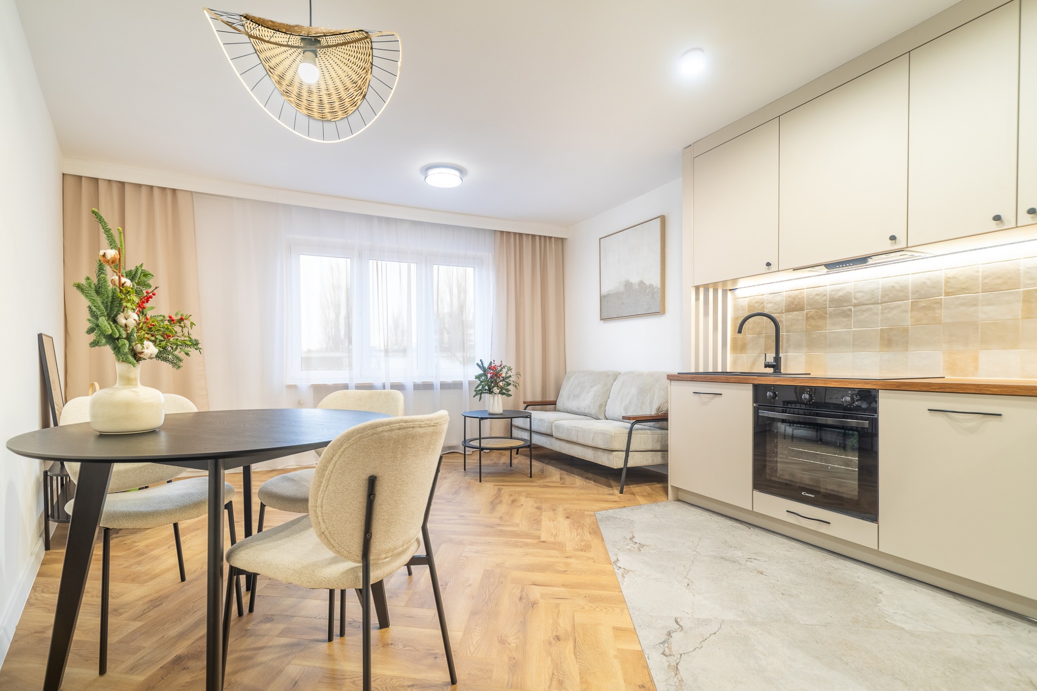 Jasne wnętrze mieszkania: stół z dekoracją, krzesła, sofa i zabudowa kuchenna z piekarnikiem. Parkiet w jodełkę, nowoczesna lampa wisząca.