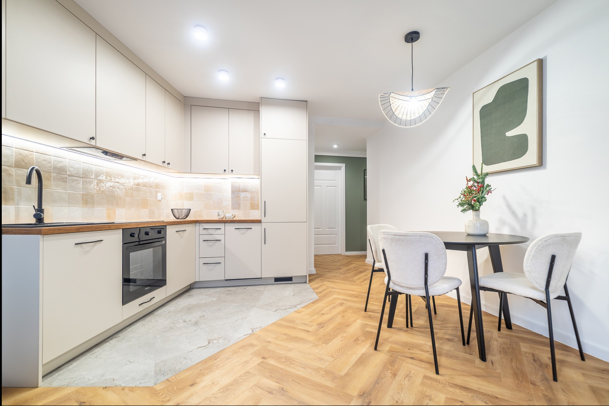 Jasna kuchnia z drewnianym blatem i parkietem w jodełkę, okrągły stół z krzesłami i dekoracją świąteczną, nowoczesne oświetlenie i minimalistyczny obraz na ścianie.