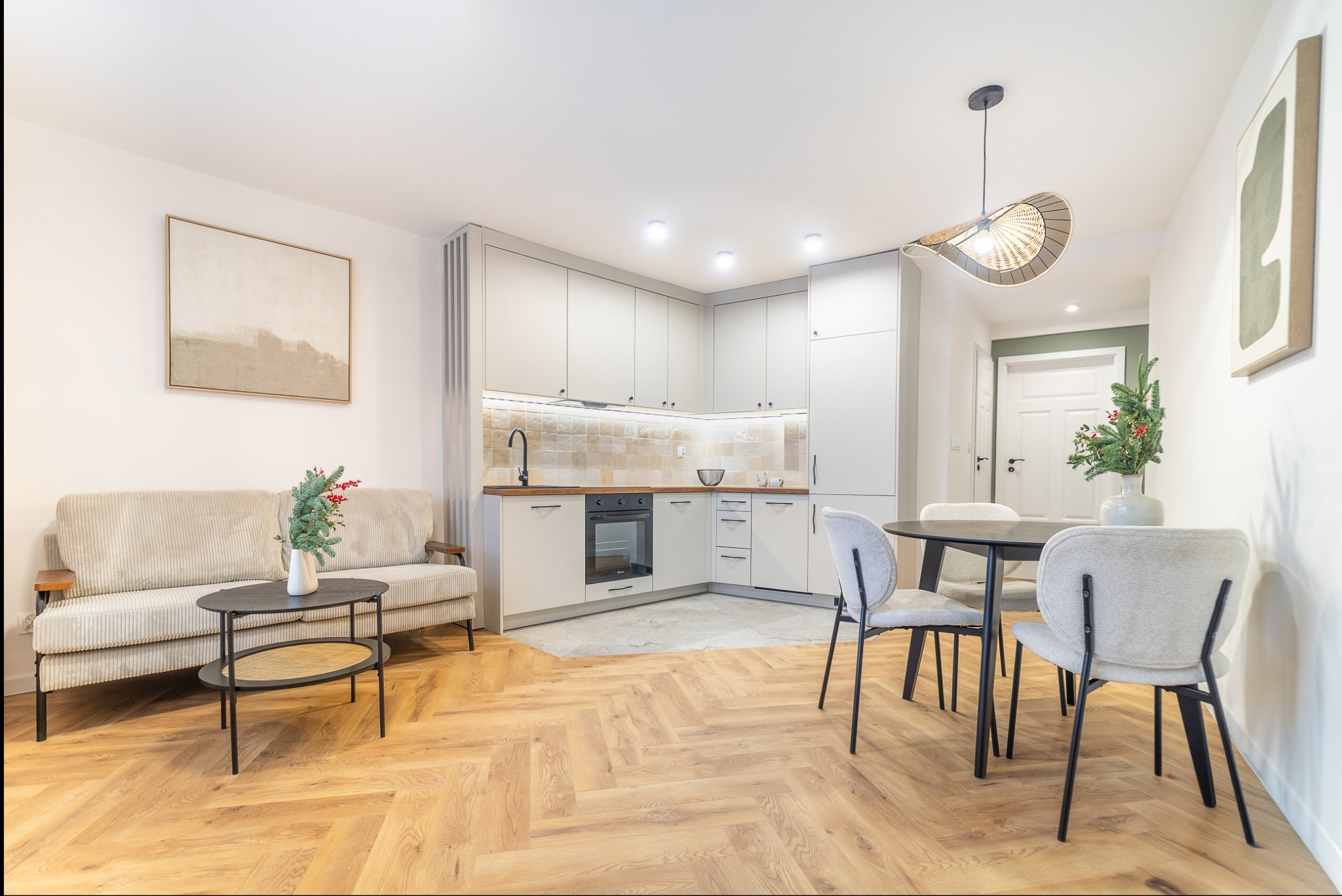 Jasne, przestronne wnętrze mieszkania z parkietem w jodełkę, beżową sofą, okrągłym stołem i nowoczesną kuchnią w odcieniach bieli i szarości. Dekoracje świąteczne dodają przytulności.