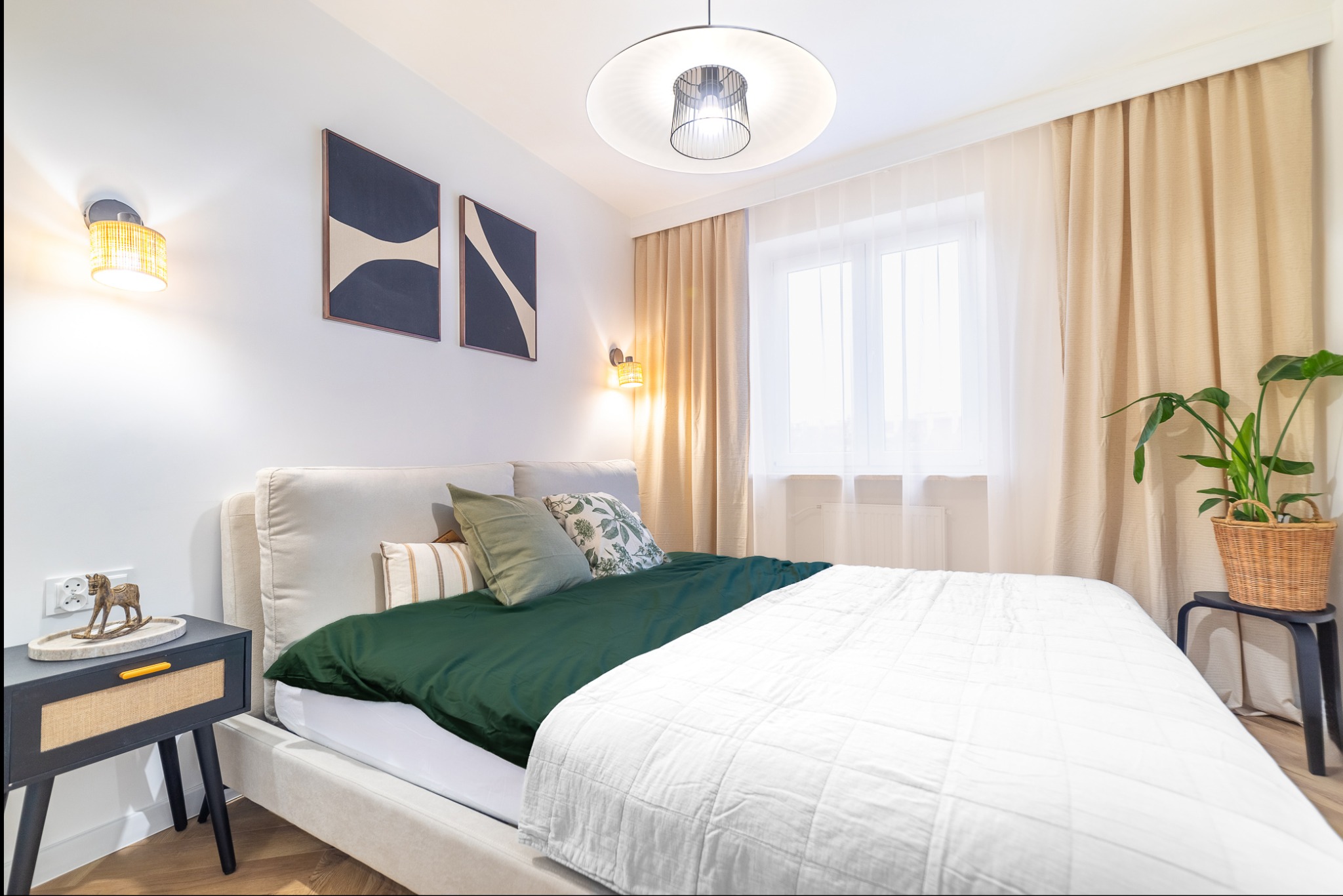 Jasna sypialnia z łóżkiem, stolikiem nocnym i rośliną doniczkową. Na ścianie abstrakcyjne obrazy i lampki. Przytulne wnętrze z beżowymi zasłonami i drewnianą podłogą.