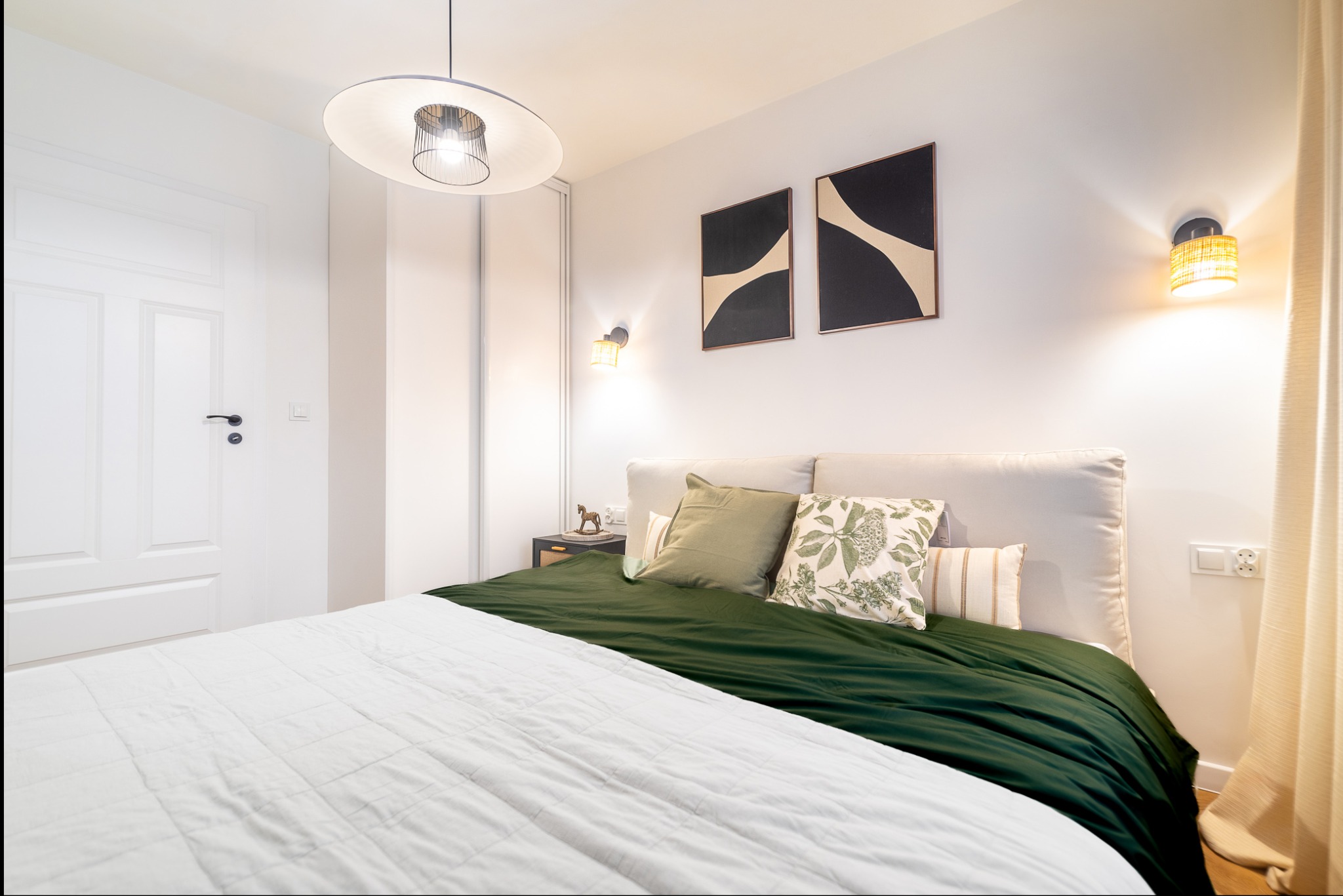 Jasna sypialnia z łóżkiem, zieloną narzutą i białymi ścianami. Nad łóżkiem wiszą abstrakcyjne obrazy w czarnych ramach i lampki nocne. Widoczna lampa sufitowa i białe drzwi.