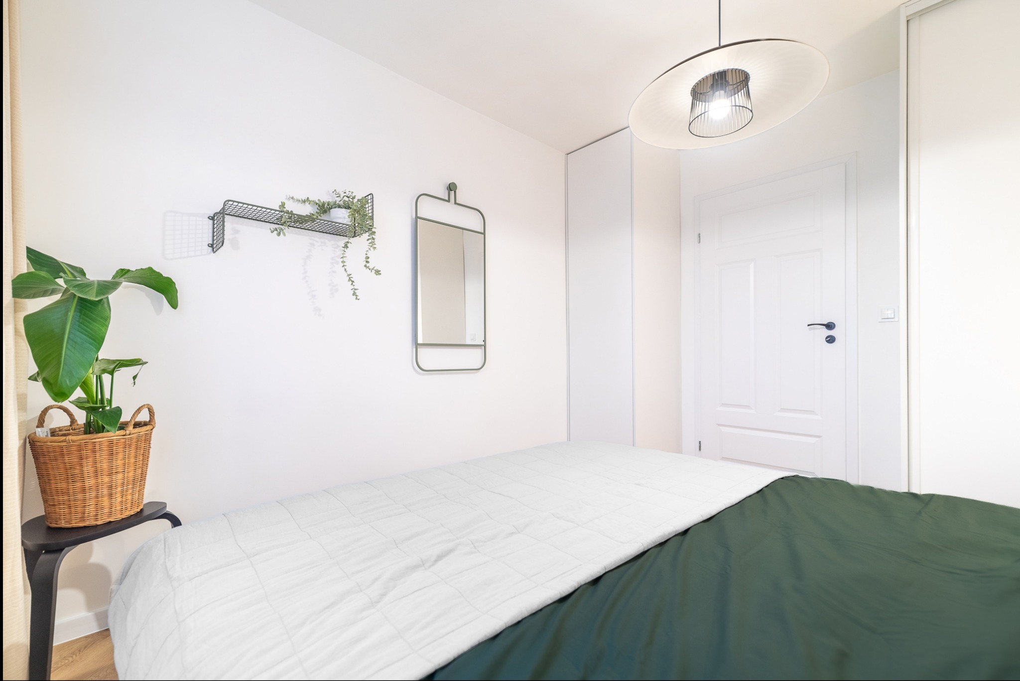 Jasna sypialnia z łóżkiem, zieloną pościelą i białym pledem. Na ścianie metalowa półka z rośliną, lustro i lampa sufitowa. Minimalistyczny design.