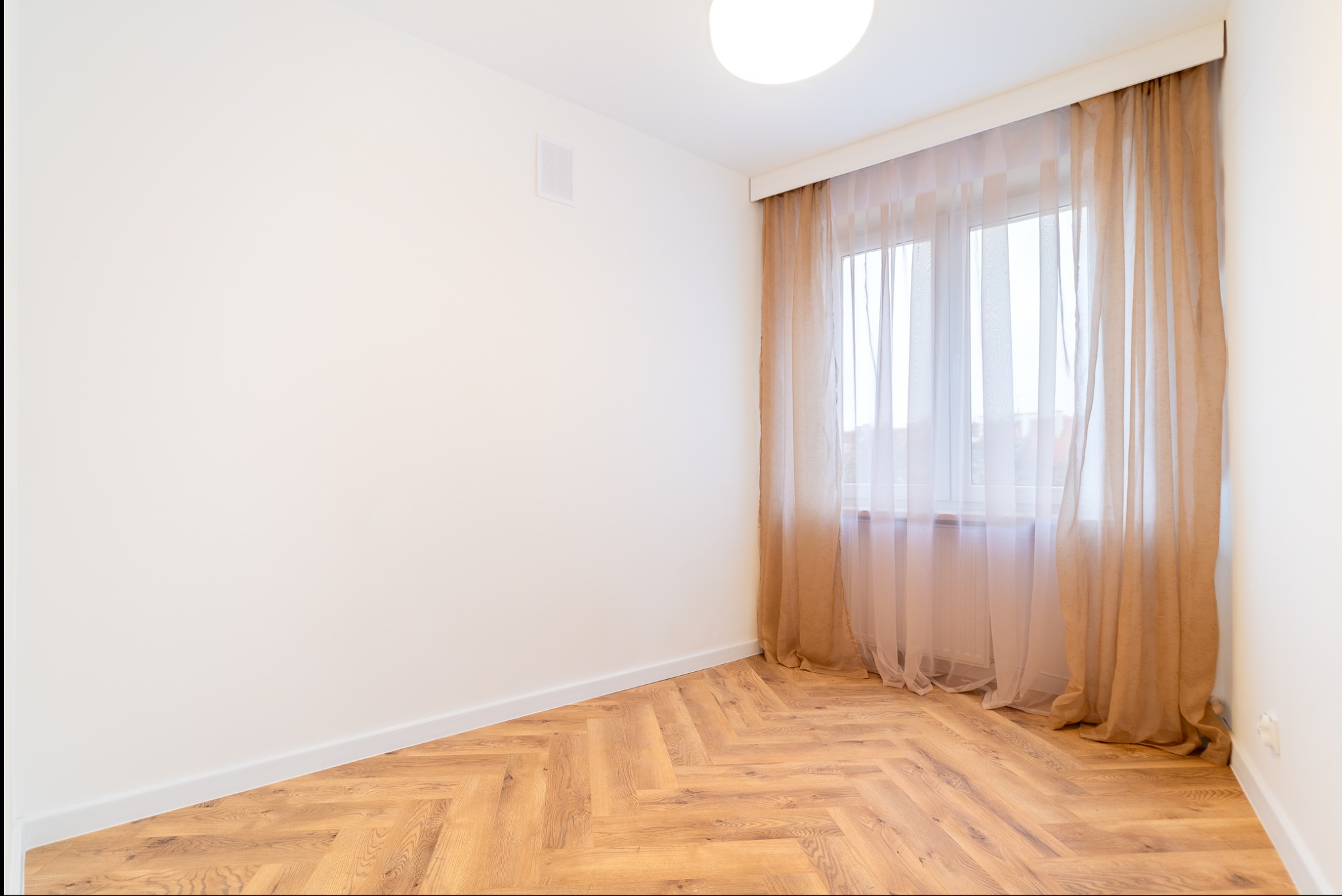 Jasne wnętrze pokoju z parkietem w jodełkę, białymi ścianami i oknem z beżowymi zasłonami. Minimalistyczny design, lampa sufitowa.