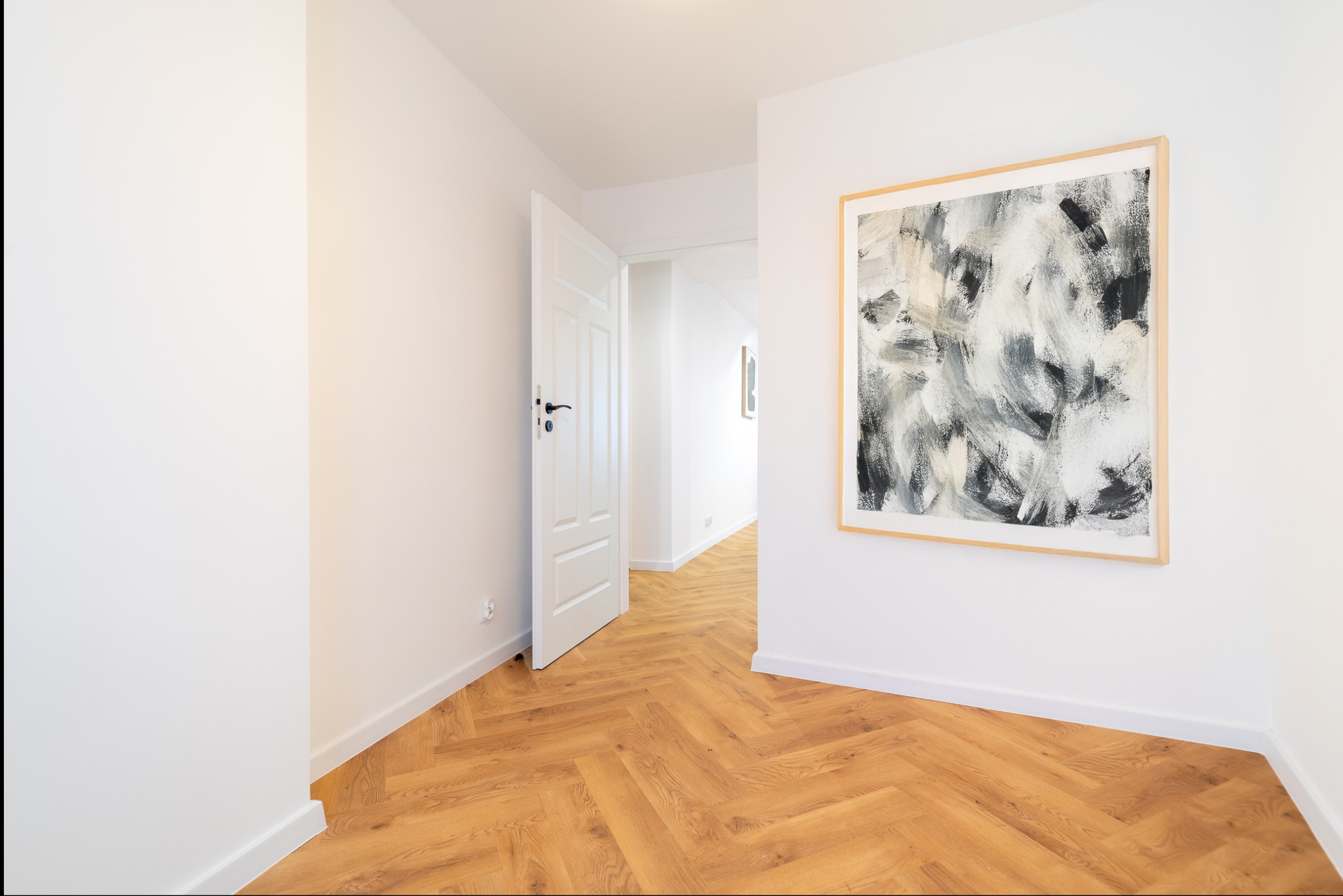 Jasne wnętrze z parkietem w jodełkę, otwartymi drzwiami i abstrakcyjnym obrazem w drewnianej ramie na białej ścianie. Minimalistyczny design.