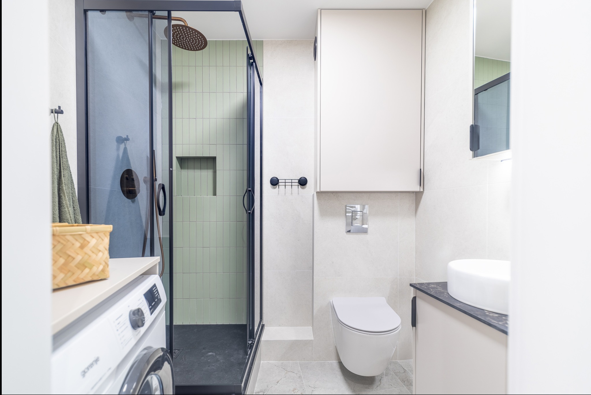 Nowoczesna łazienka po remoncie: kabina prysznicowa z zielonymi płytkami, wisząca toaleta, umywalka na blacie i pralka. Minimalistyczny design, jasne kolory.