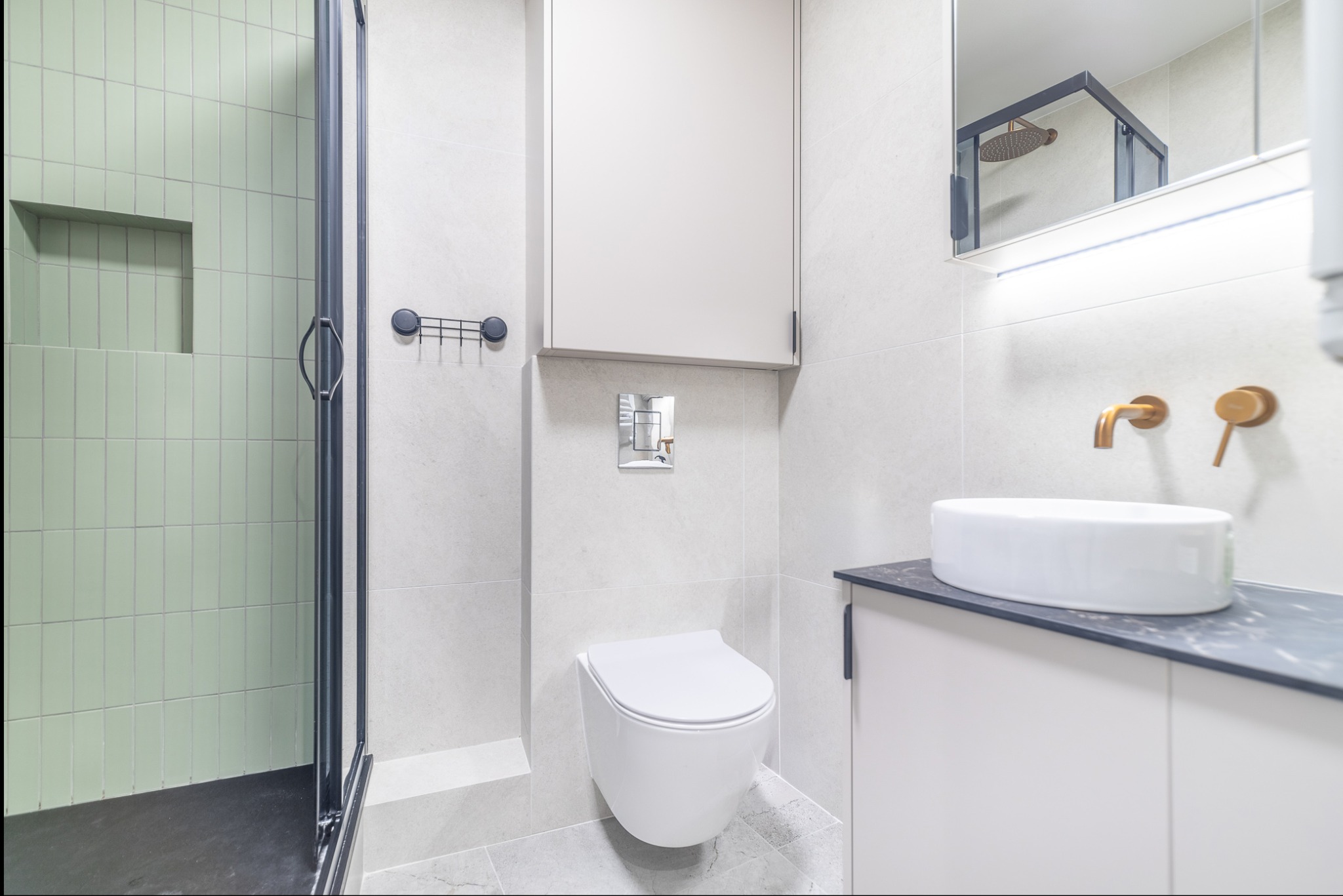 Nowoczesna łazienka z kabiną prysznicową z zielonymi płytkami, podwieszaną toaletą i umywalką nablatową na szafce z blatem imitującym kamień.