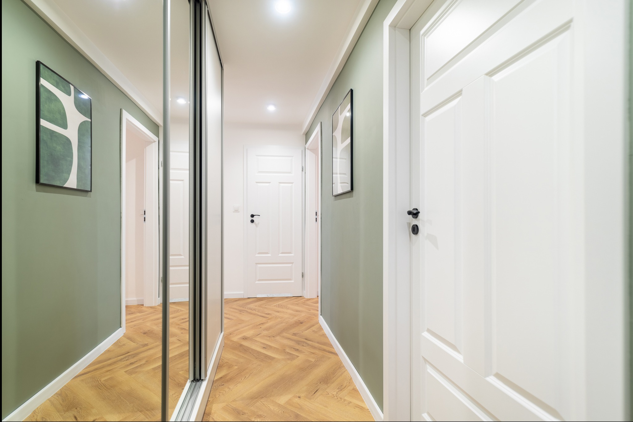 Nowoczesny korytarz z parkietem w jodełkę, szafą z lustrzanymi drzwiami i zielonymi ścianami. Minimalistyczny design, białe drzwi i oświetlenie punktowe.