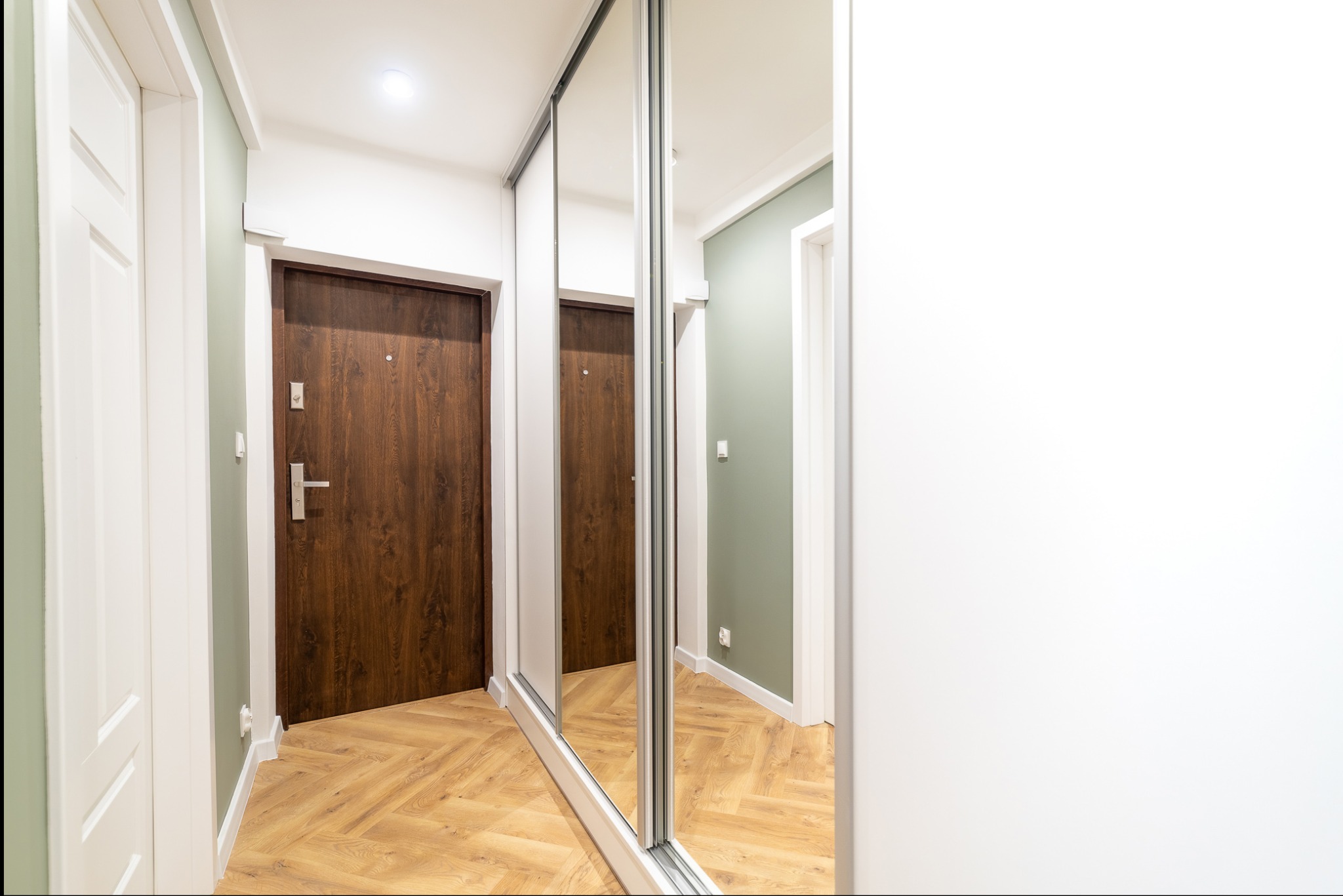 Nowoczesny przedpokój z parkietem w jodełkę, lustrzaną szafą przesuwną i dwiema parami drzwi w odcieniach bieli i brązu. Ściany białe i oliwkowe.