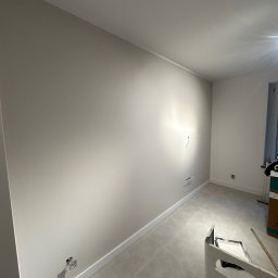 NowaPrzestrzeń - Jasne wnętrze po malowaniu ścian na beżowo, z widocznymi gniazdkami elektrycznymi i listwami przypodłogowymi. Na podłodze szare płytki. Widoczny kuwet z farbą.