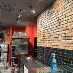 NowaPrzestrzeń - Wykończona ściana z cegły i czarnych płytek w lokalu gastronomicznym. Widoczny sprzęt kuchenny i środki czystości. Oświetlenie sufitowe.