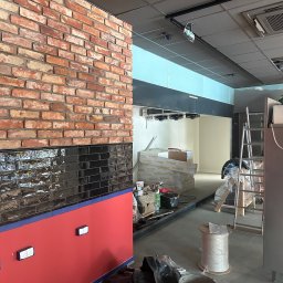 NowaPrzestrzeń - Wnętrze w trakcie prac wykończeniowych: ściana z cegły i czarnych płytek, czerwona dolna część ściany, widoczne materiały budowlane i drabina. Oświetlenie sufitowe.