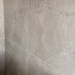 NowaPrzestrzeń - Ściana wyłożona szarymi, matowymi płytkami imitującymi kamień z delikatną fakturą i jasnymi fugami. Widok z bliska, ukazujący precyzję ułożenia.