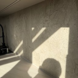 NowaPrzestrzeń - Beżowa ściana z marmurowym wzorem w kuchni, oświetlona promieniami słońca tworzącymi geometryczne cienie. Widoczny fragment blatu i czarna bateria.