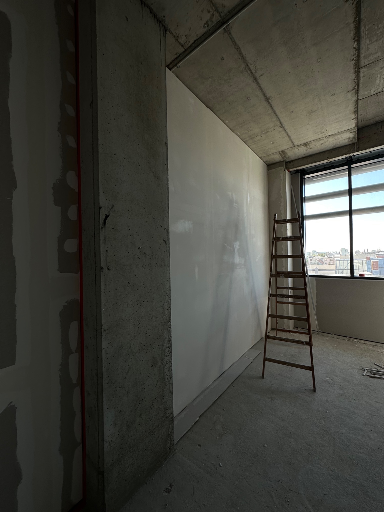 Wnętrze w trakcie prac wykończeniowych: gładź na ścianie, surowy betonowy strop, drabina oparta o ścianę, widok z okna na miasto.