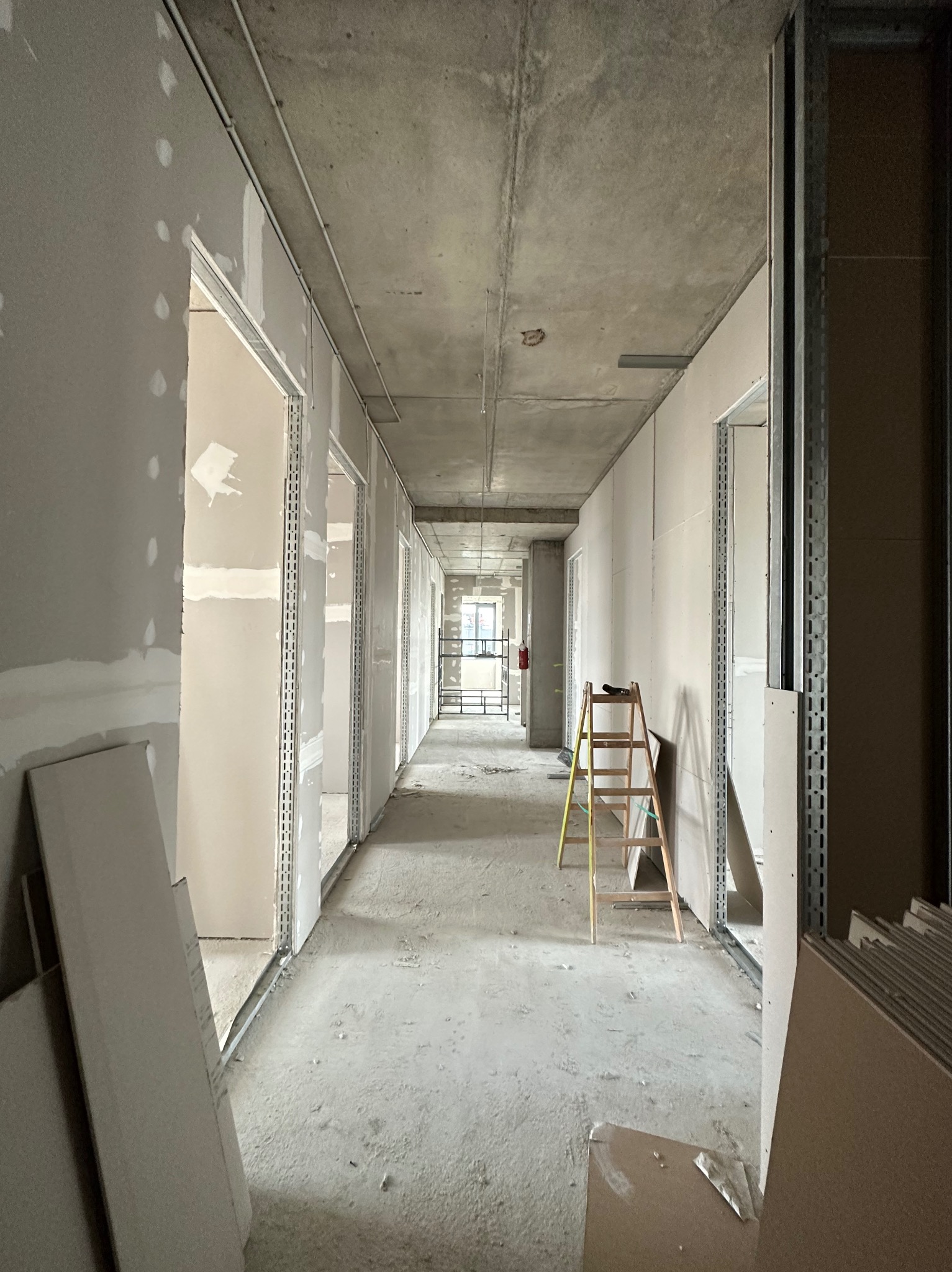 Korytarz w trakcie szpachlowania i montażu płyt gipsowo-kartonowych. Widoczna drabina i profile. Surowy betonowy sufit. Długi kadr z perspektywą.