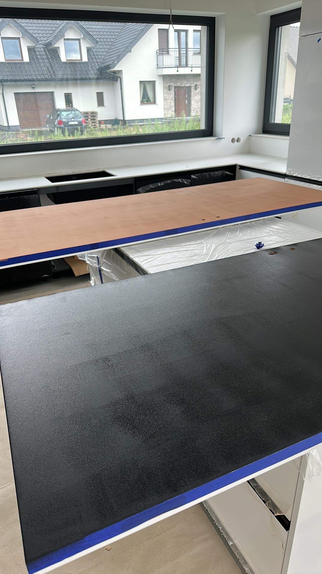 Częściowo zamontowany, czarny blat kuchenny z zabezpieczonymi krawędziami, widok na okno z domem w tle. Etap montażu blatu w nowej kuchni.